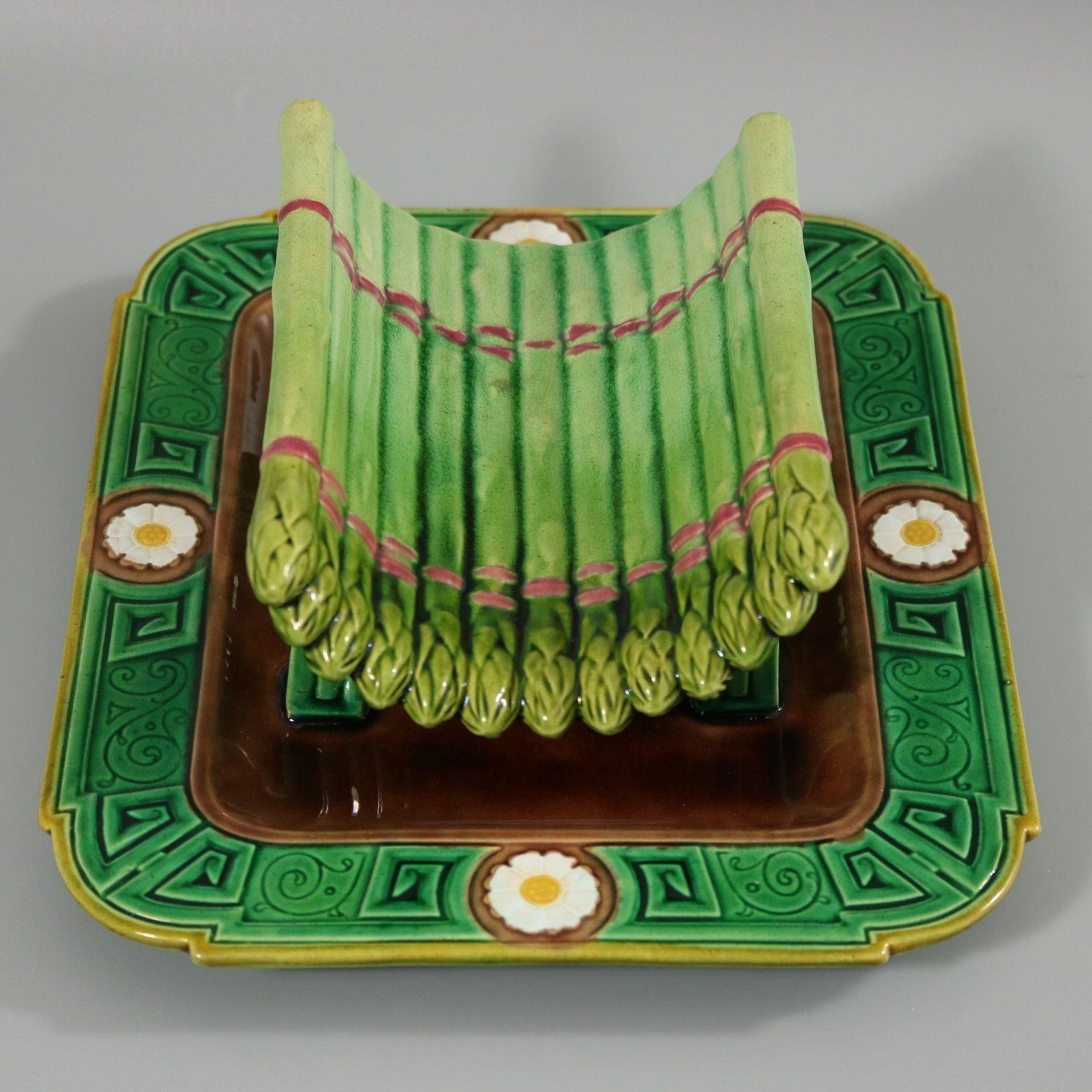 Minton Majolica asparagus drainer which features asparagus tips bound together, sat on a rectangular tray. Flowers and geometric patterns to the border. Colouration: green, brown, white, are predominant. The piece bears maker's marks for the Minton