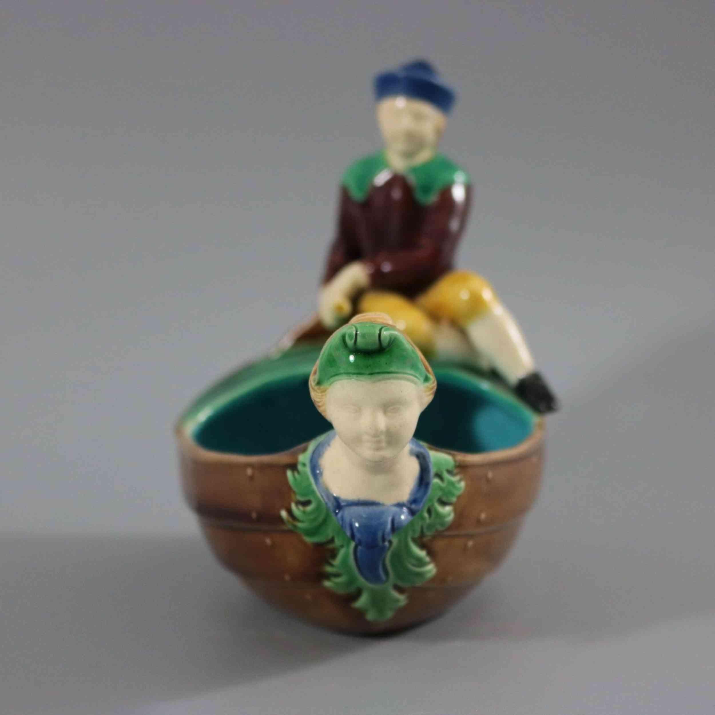 Minton Majolica dish which features a boat with a boy sitting on the back, steering the rudder. A woman's bust figurehead at the front of the boat. Colouration: brown, green, turquoise, are predominant. The piece bears maker's marks for the Minton