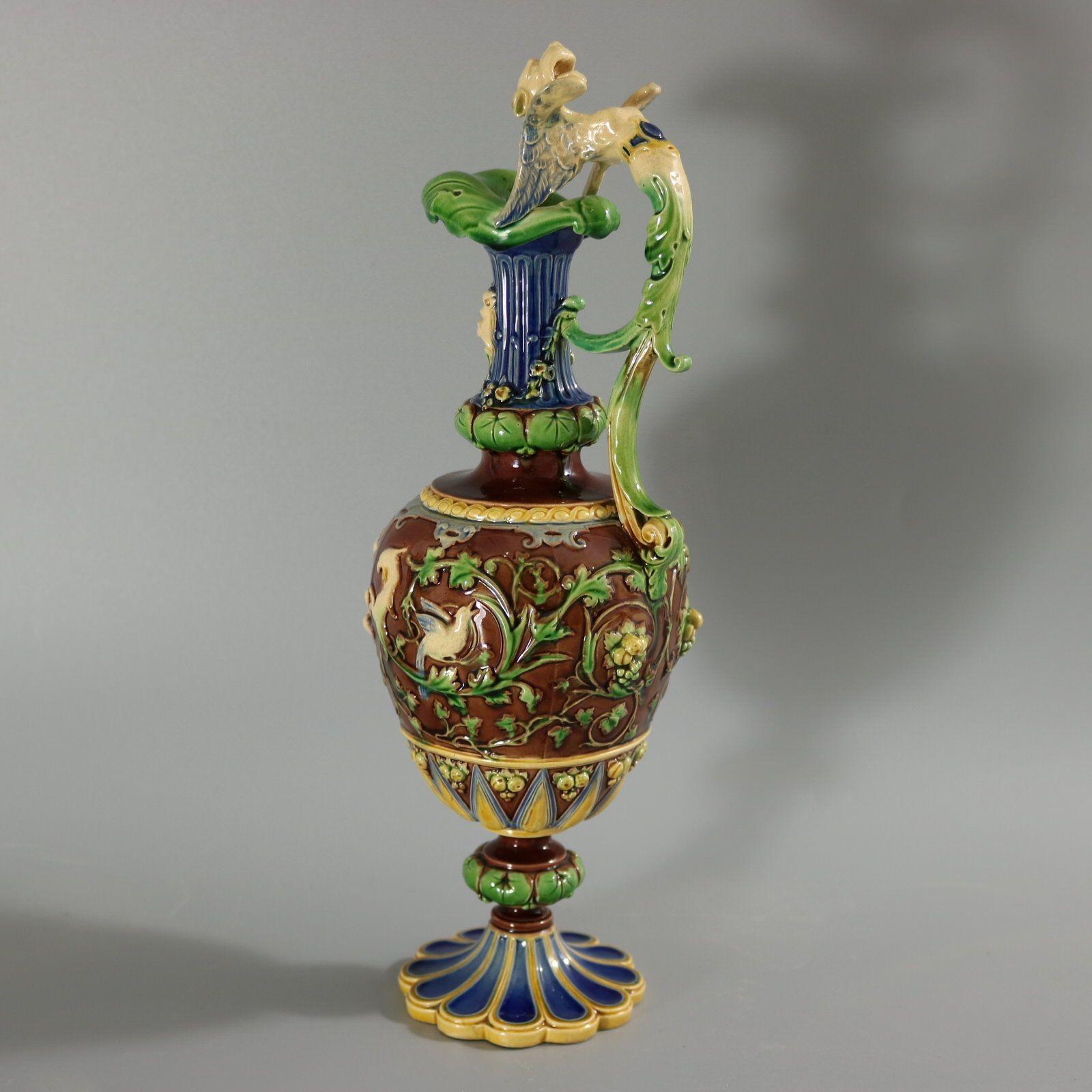 Minton Majolica ewer with a mythological theme which features putti, birds and vine fruits. The ewer has a tapered neck, with mask head and Sphinx handle. Sits on circular fluted foot. Colouration: brown, green, blue, are predominant. Marks include