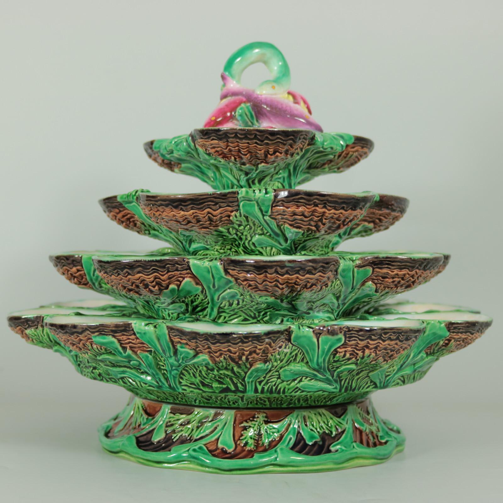 Minton Majolica oyster stand which features four tiers of open oyster shells, laid on seaweed. A fish and eel handle at the top. Rotates on the base. Colouration: green, white, brown, are predominant. Bears a monogram mark, 'CB'.