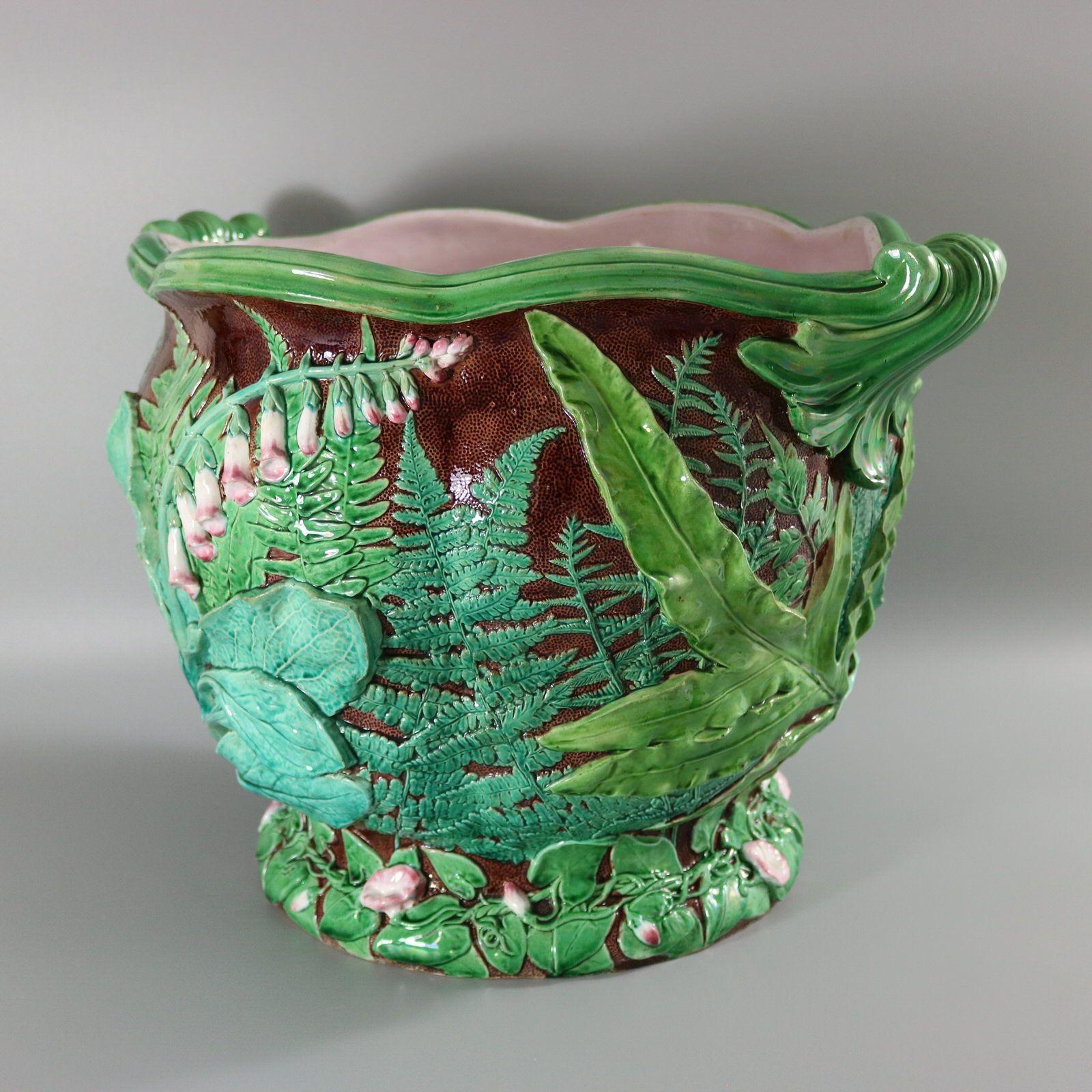 Minton Majolica jardinière which features foxgloves, ferns and lush, leafy foliage on brown stipple ground. Two green scroll handles on the shaped rim. Brown ground version. Colouration: green, brown, pink, are predominant. Bears a pattern number,