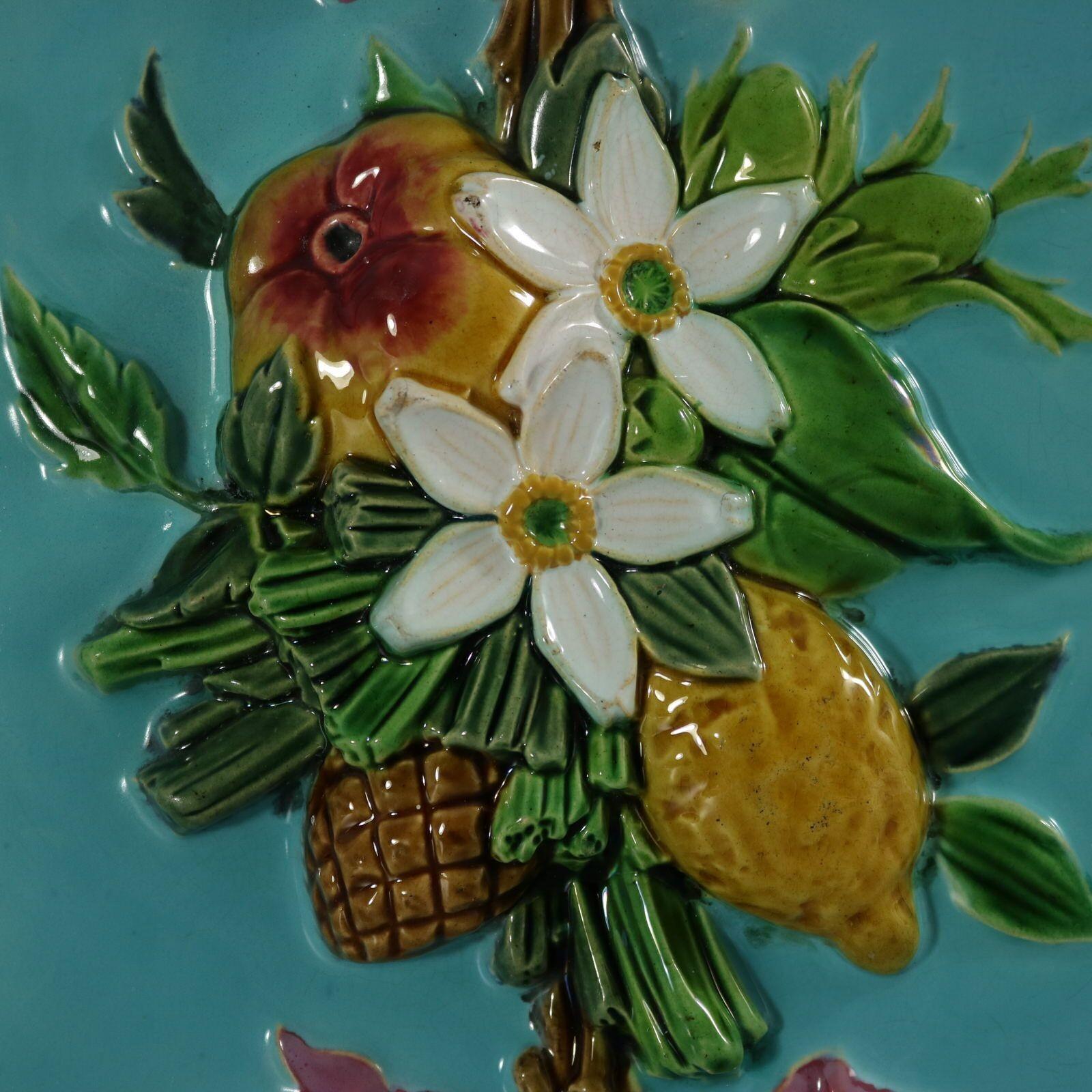 Minton Majolica tile which features an assemblage of fruit, flowers and leaves, including an apple, a lemon and a pinecone. Ribbons on either side. Colouration: turquoise, green, white, are predominant. The piece bears maker's marks for the Minton
