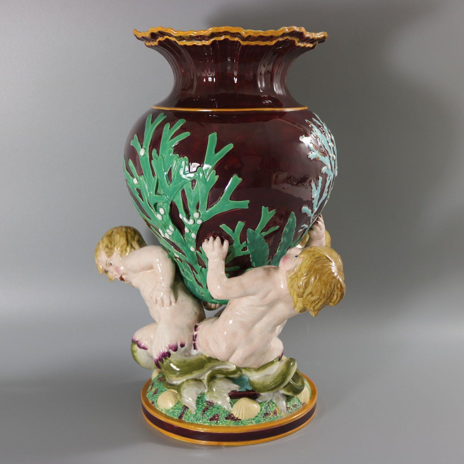Minton Majolica vase which features three merboys supporting a vessel adorned with seaweeds. The rim of the vase is modelled to depict breaking waves. Dark maroon ground version. Colouration: dark maroon, green, pink, are predominant.