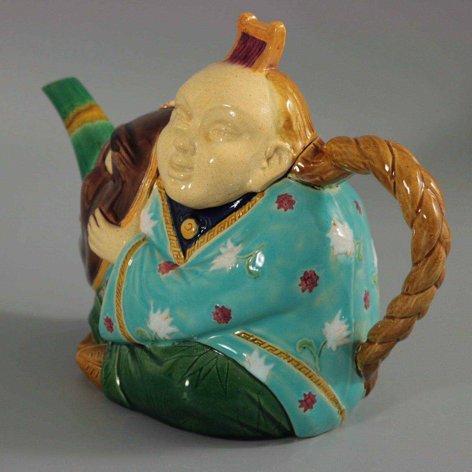 Minton Majolica teapot which features an oriental man, dressed in a floral robe, holding a Noh mask. His hair forms the handle. Colouration: turquoise, brown, green, are predominant. The piece bears maker's marks for the Minton pottery. Bears a