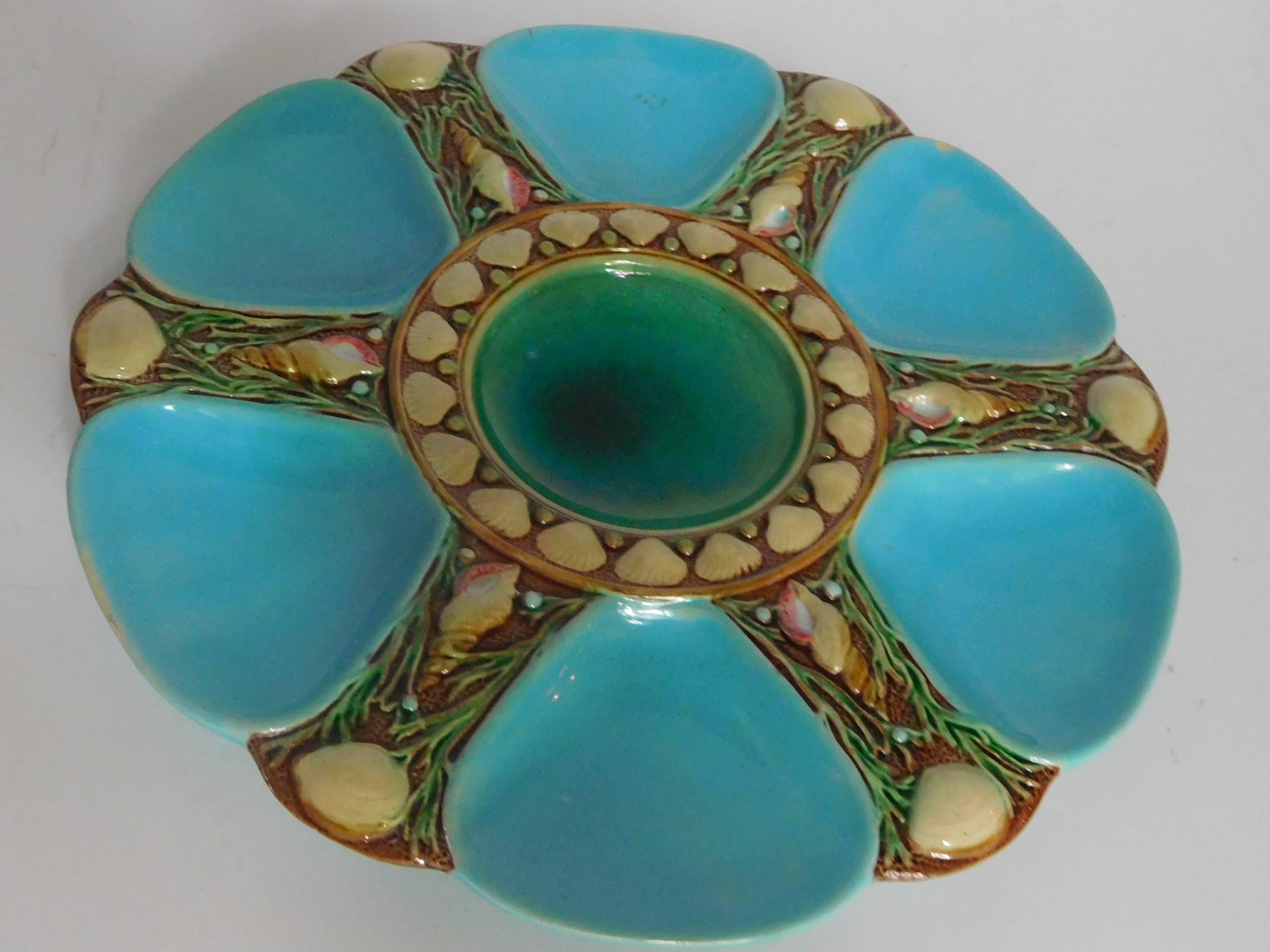 This colorful oyster plate has six turquoise-colored wells shaped like shells on a bed of little white shells and dark seaweeds. In the center is a sauce well in deep ocean green surrounded by a parade of pearls and shells. This pattern, sold in