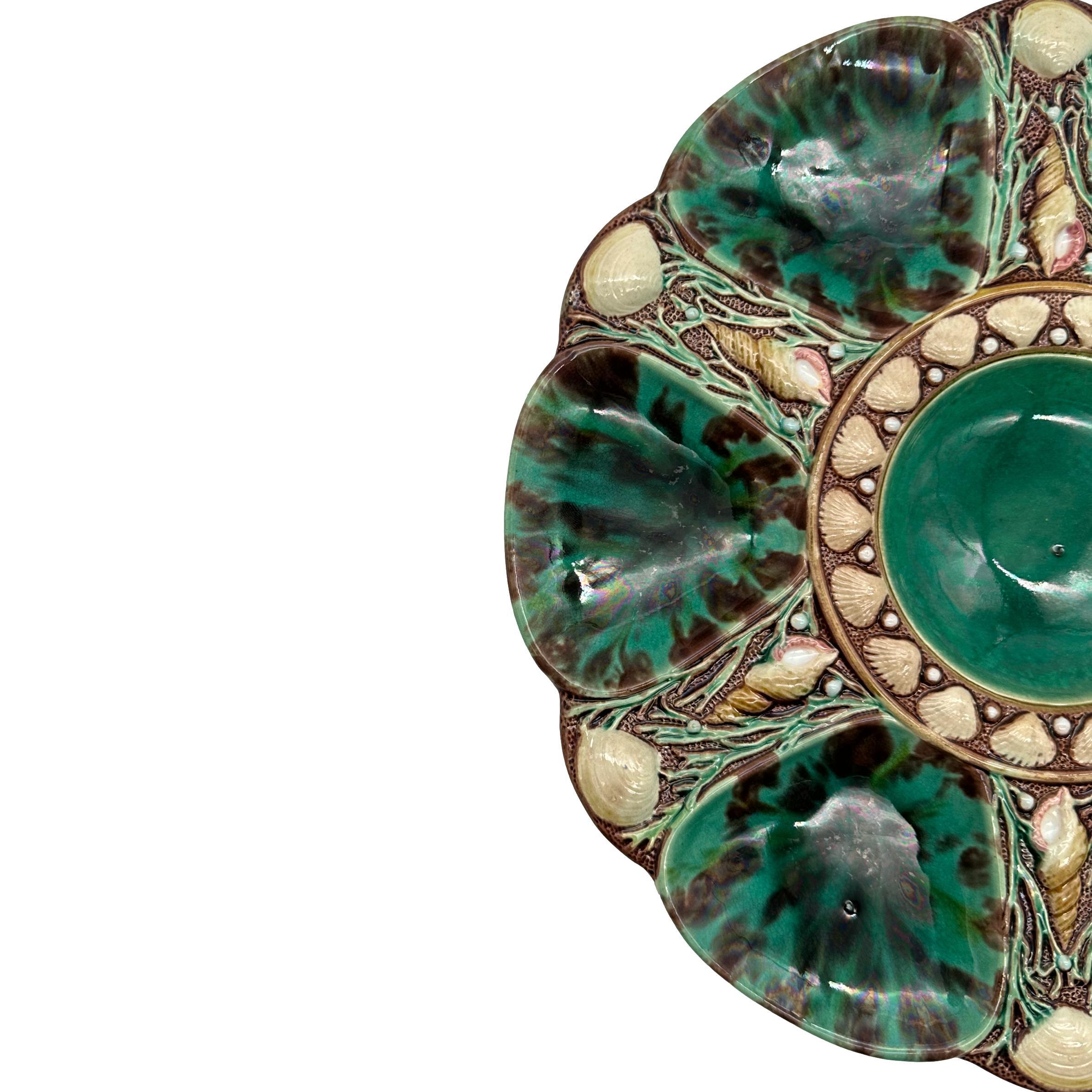 Minton Majolica Oyster Plate, the relief-molded dish with six wells glazed in green and brown mottled leopard spots, each well separated by shells and seaweed, the center well glazed in green and banded with shells, the reverse with impressed and