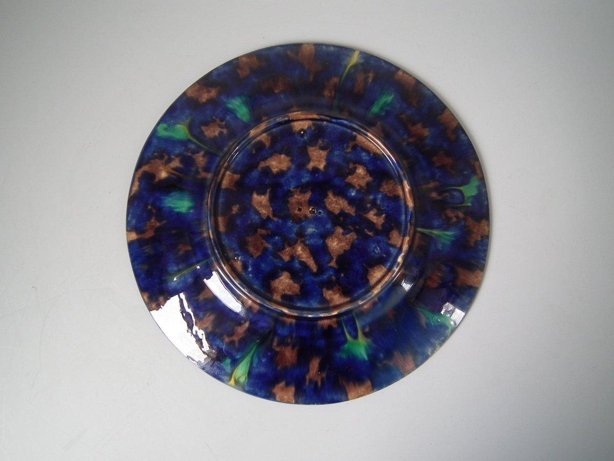 Minton Majolica plate which features intaglio grape and vine pattern to centre, six mottled cartouches to the outer edge. Colouration: green, cobalt blue, ochre, are predominant. Marks include a factory specific date cipher for the year 1858.
 