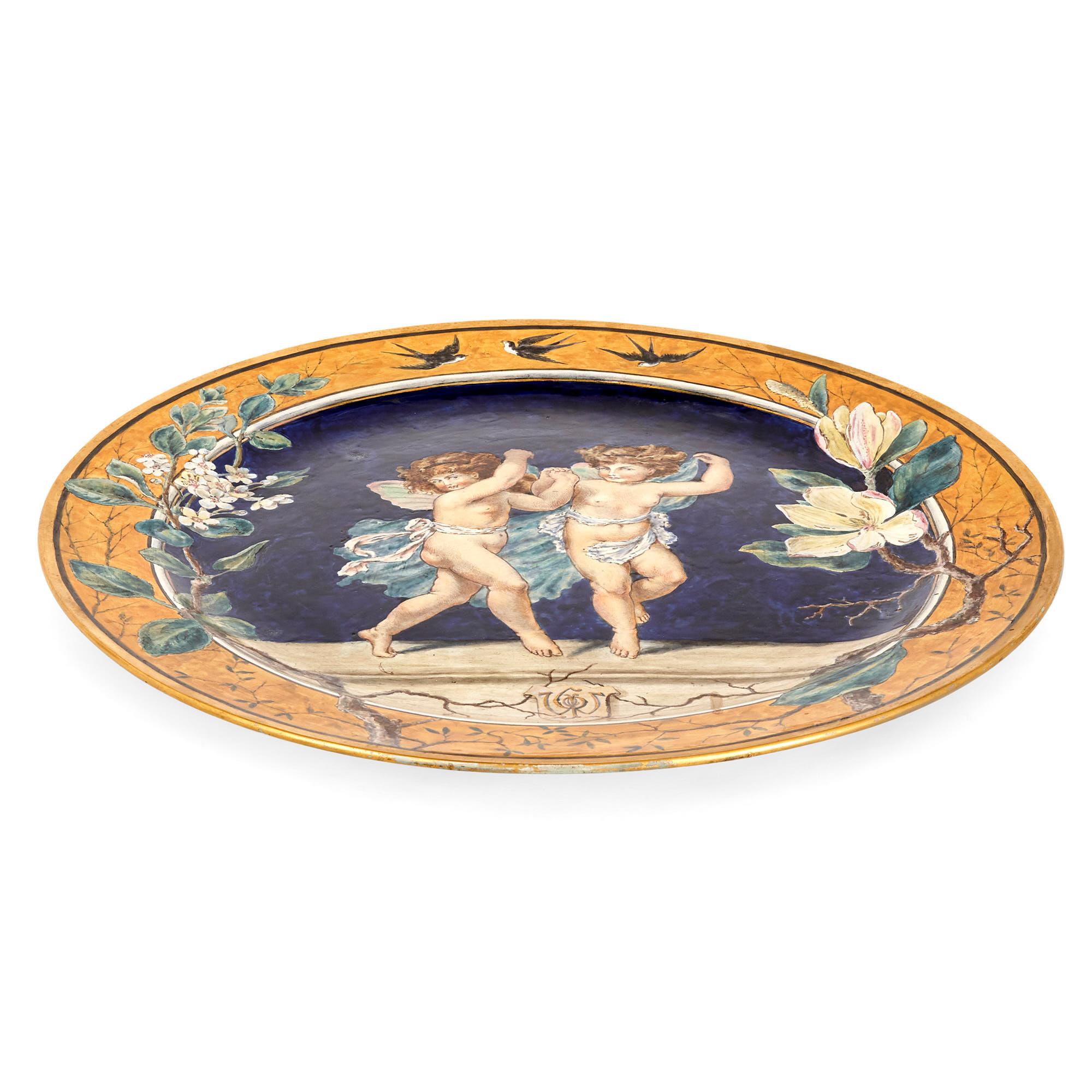 This beautiful large plate, its diameter is over 50 cm, was created by the English firm Minton and is crafted from tin-glazed ceramic, also known as Majolica. The surface of the plate is decorated with two cherubim set against a rich blue backdrop.