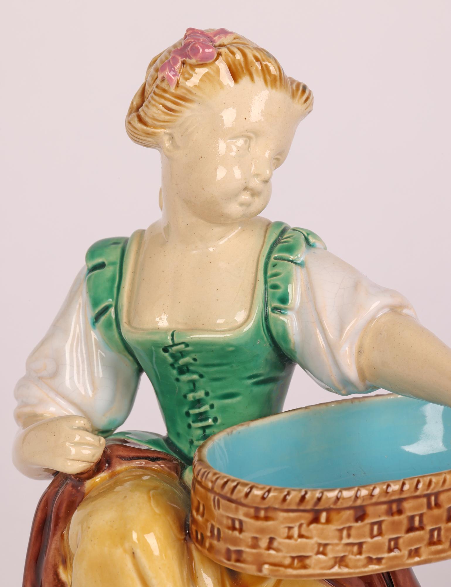 A stunning Minton majolica pottery figure depicting a girl harvester figurine dating from 1864. The figure sits raised on a round pedestal base and holds a large oval shaped basket under one arm. The figure is finely detailed and hand painted in