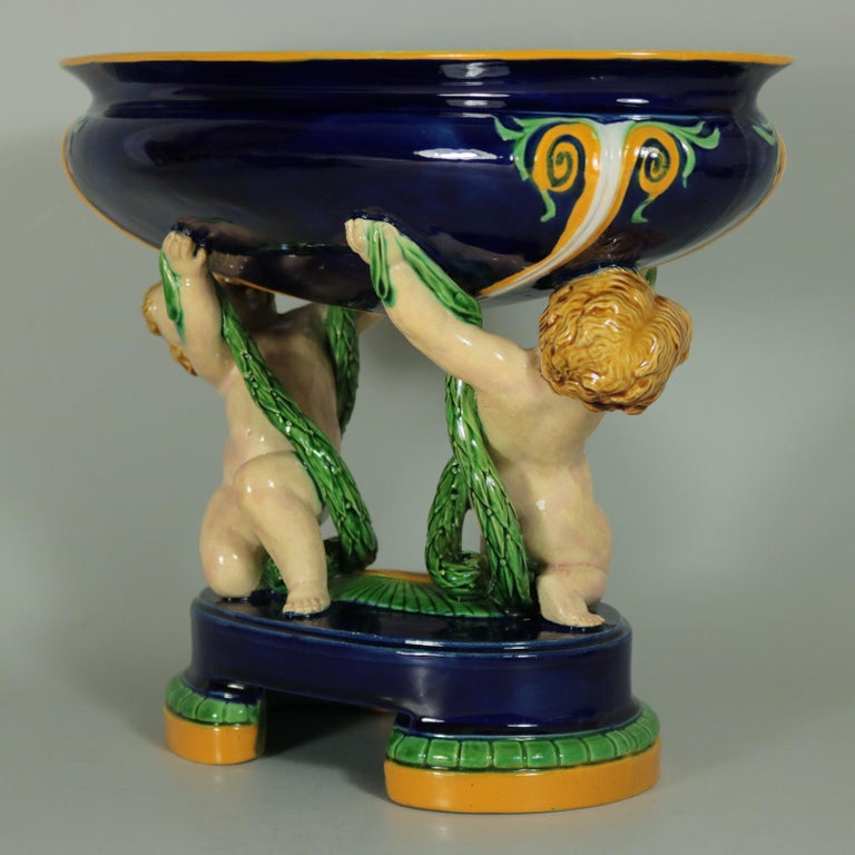 Minton Majolica figural bowl which features putti holding leafy garlands, supporting the bowl. Colouration: cobalt blue, green, cream, are predominant. The piece bears maker's marks for the Minton pottery. Bears a pattern number, '1647'. Marks