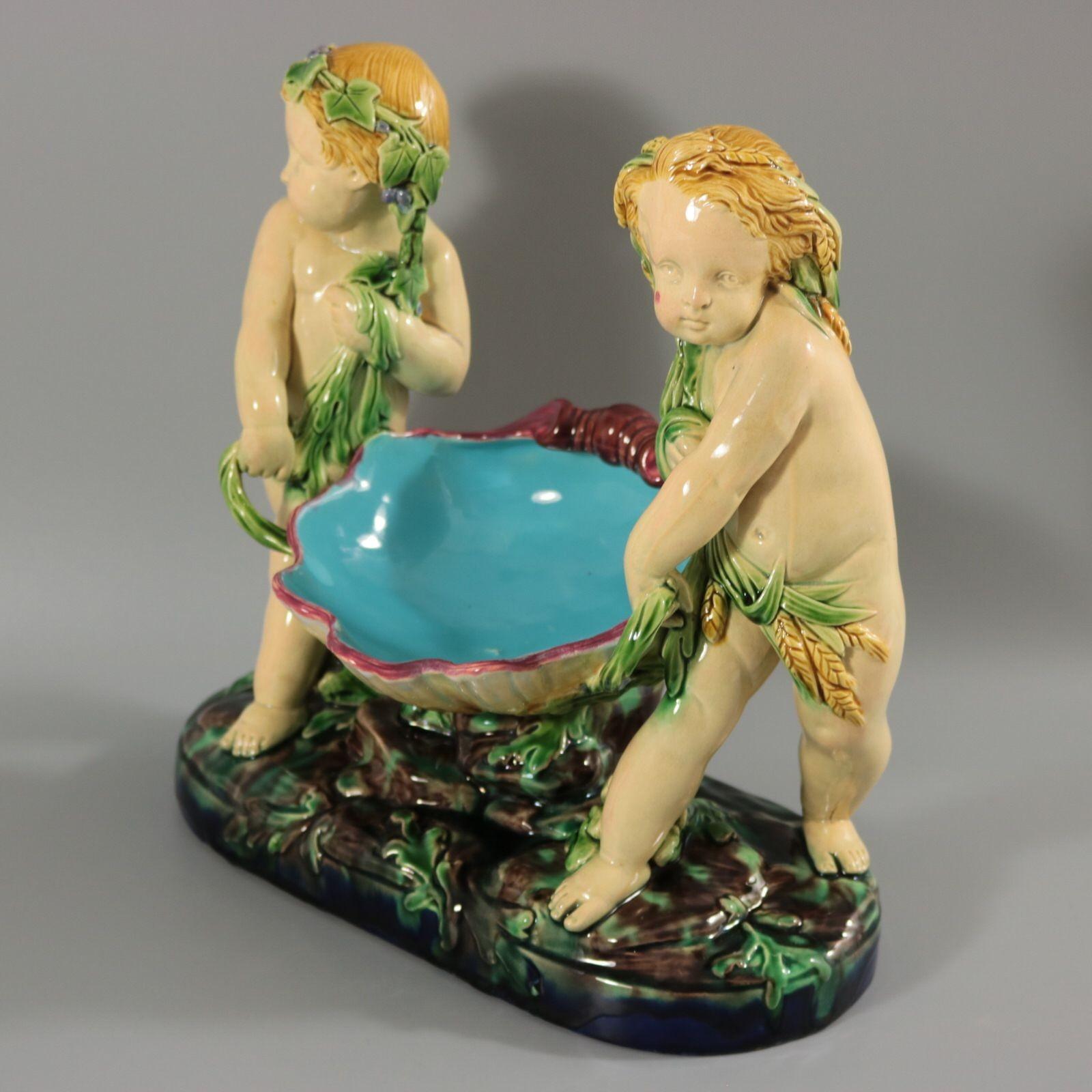 Minton Majolica figural which features two putti carrying a shell. One draped in grapes, the other draped in wheat. Colouration: green, turquoise, cream, are predominant. The piece bears maker's marks for the Minton pottery. Bears a pattern number,