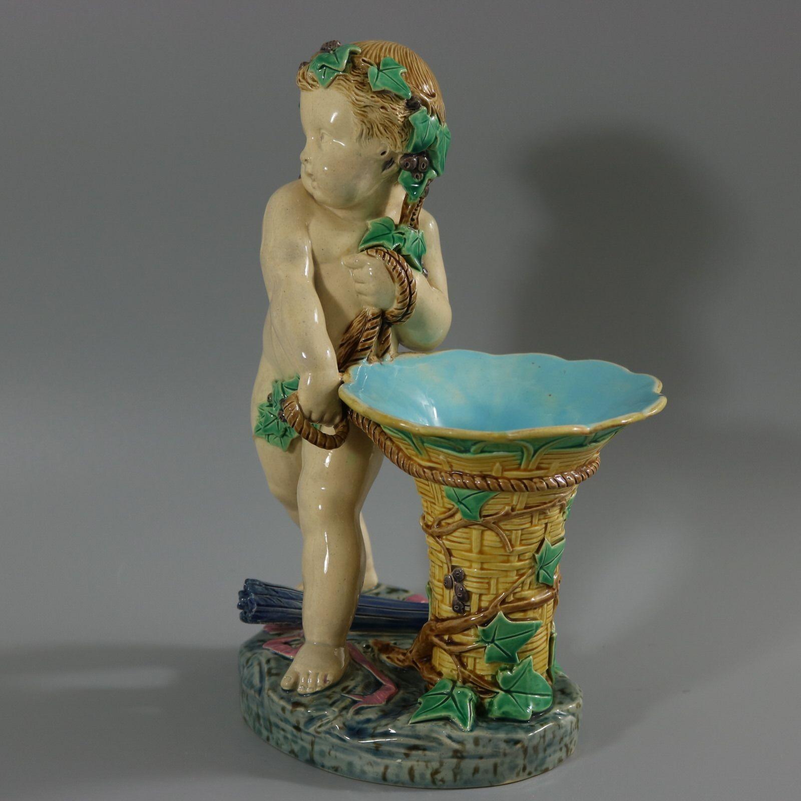 Minton Majolica figural vase which features a putti holding a basket, with a quiver of arrows between his feet. Colouration: cream, yellow, blue, are predominant. The piece bears maker's marks for the Minton pottery. Bears a pattern number, '405'.
