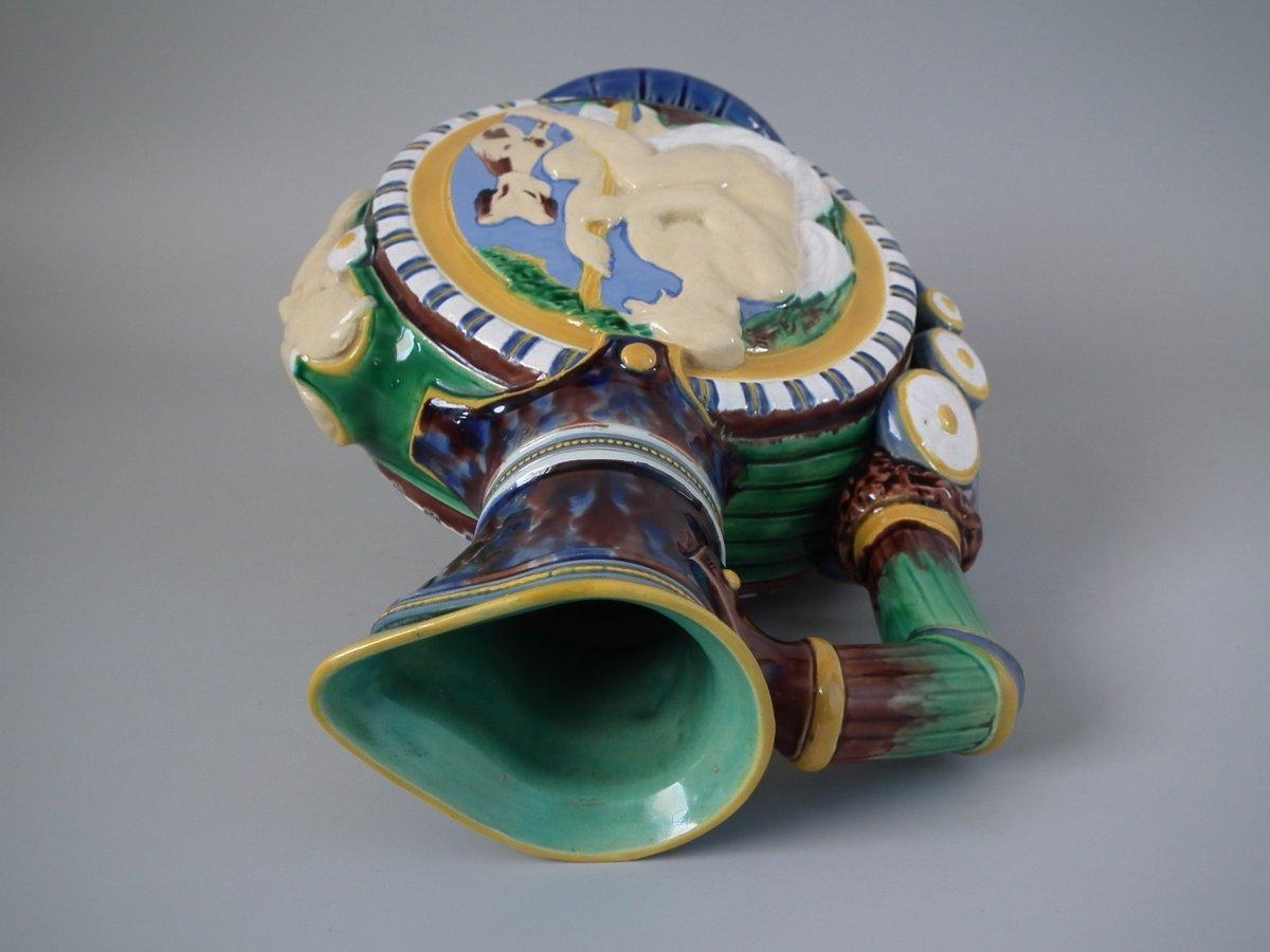 Minton Majolica jug or pitcher which features circular panels to the sides, depicting classical figures with dogs. (Diana, the huntress on one side.) Grotesque mask to the front. Coloration: Green, blue, ochre, are predominant.