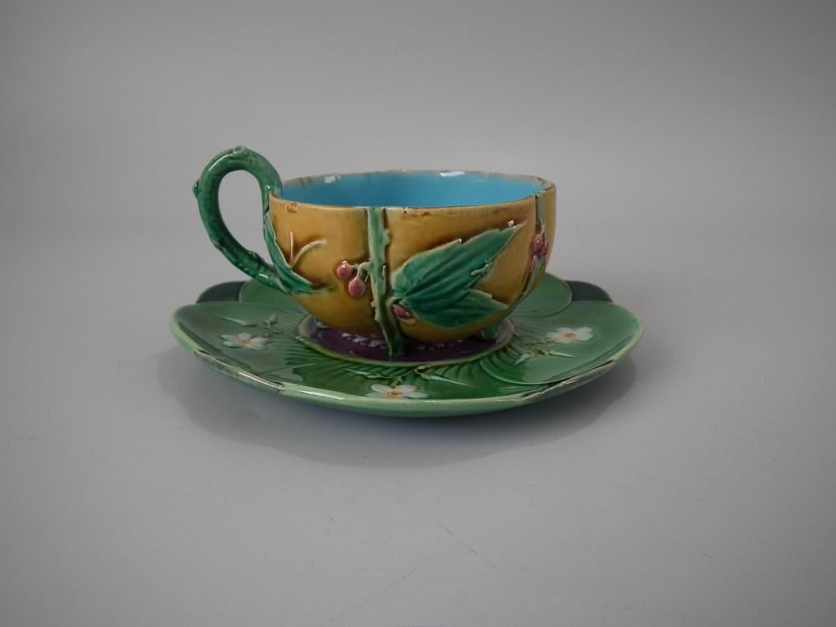 Minton Majolica cup and saucer which features lily pads, leaves and blossom. Coloration: green, ochre, white, are predominant. The piece bears maker's marks for the Minton pottery. Bears a pattern number, '1349'. Marks include a factory specific