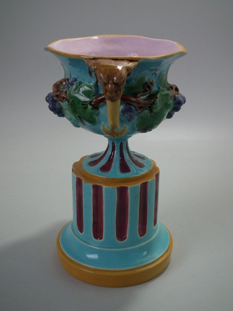 Minton Majolica vase which features grapes and vine leaves, two lion head handles and a (fixed) pedestal base. Colouration: turquoise, pink, ochre, are predominant. The piece bears maker's marks for the Minton pottery. Bears a pattern number, '500'.