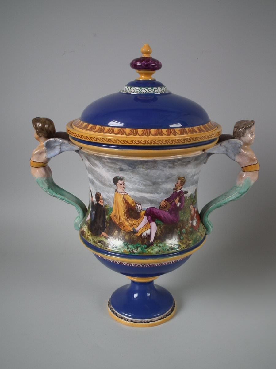 Rare Minton Tin-Glazed Majolica Pictorial lidded vase with cupid handles. Features a pictorial scene of people in a country landscape. The vase was painted by J D Rochfort - an artist who painted Minton pottery, active 1860s-1870s, who took up