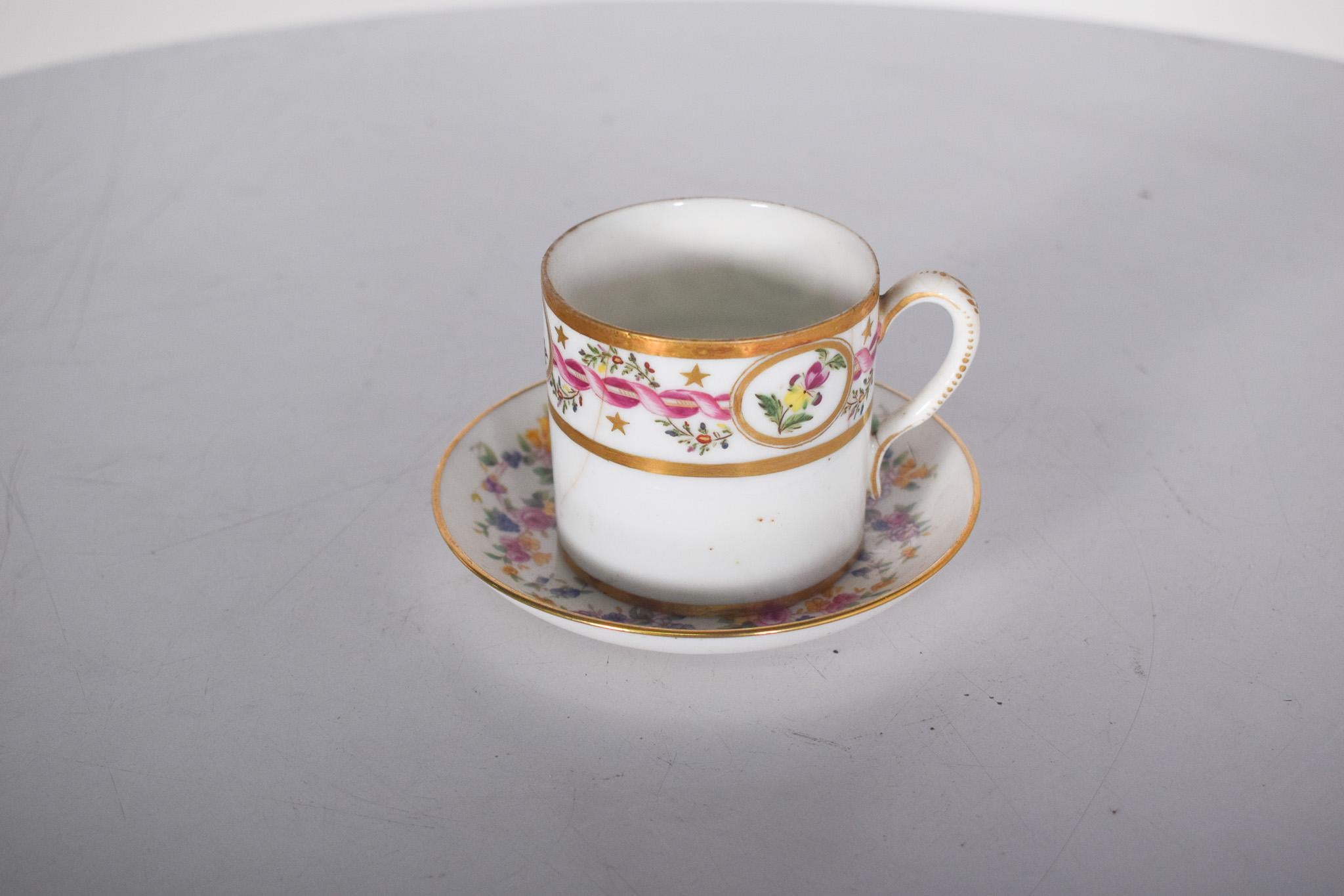 Taza y platillo de porcelana inglesa Mintons. Decorada con un delicado motivo floral y detalles dorados, la taza tiene una forma cilíndrica clásica, adornada con un borde dorado que recorre tanto la parte superior como la base. La decoración está