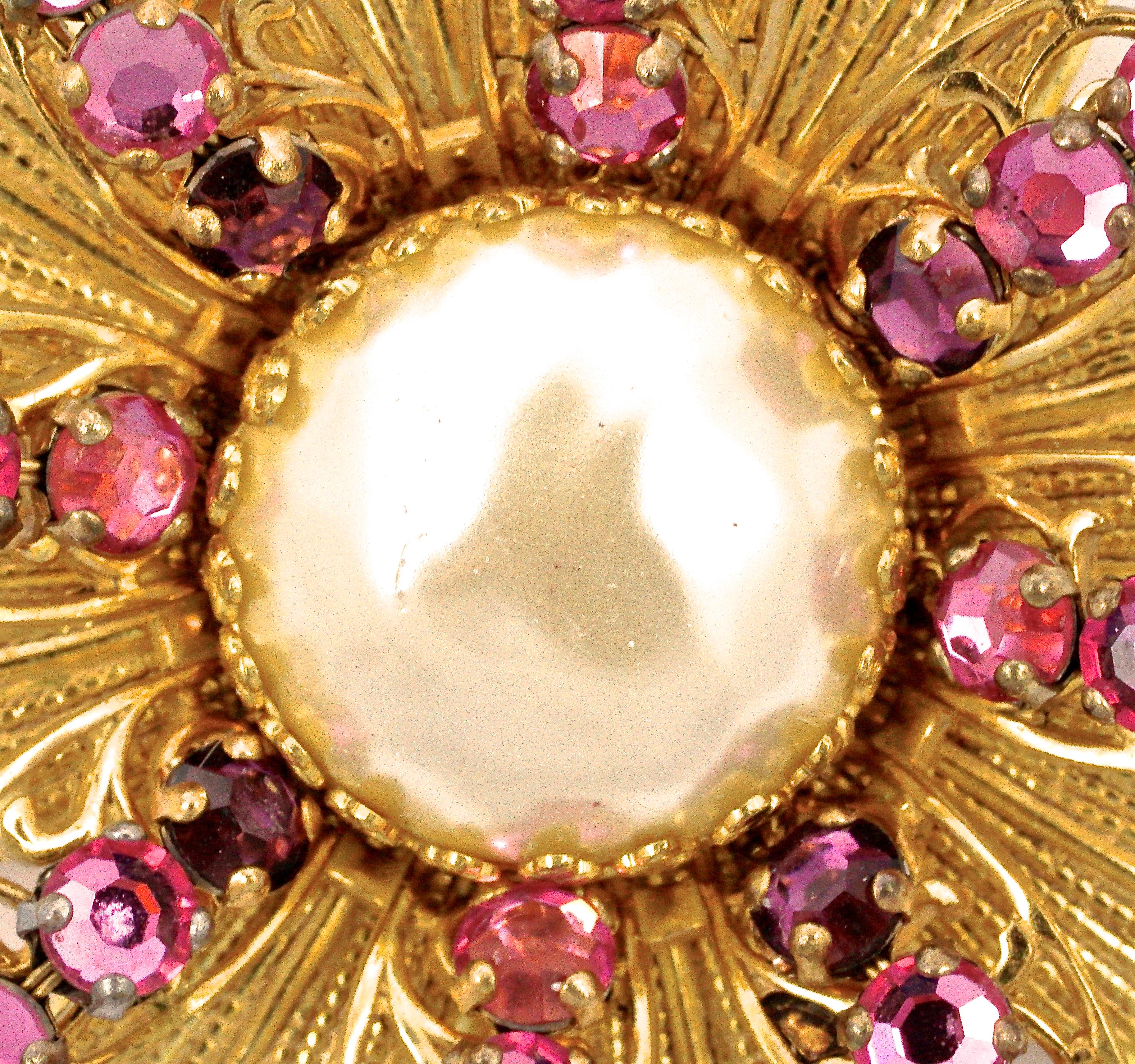 
Beautiful Miriam Haskell gold tone flower brooch featuring a large faux baroque pearl, and wired rose montee pink and purple rhinestones. The pearl and rhinestone petals are layered on to golden textured petals, and the back has a filigree frame