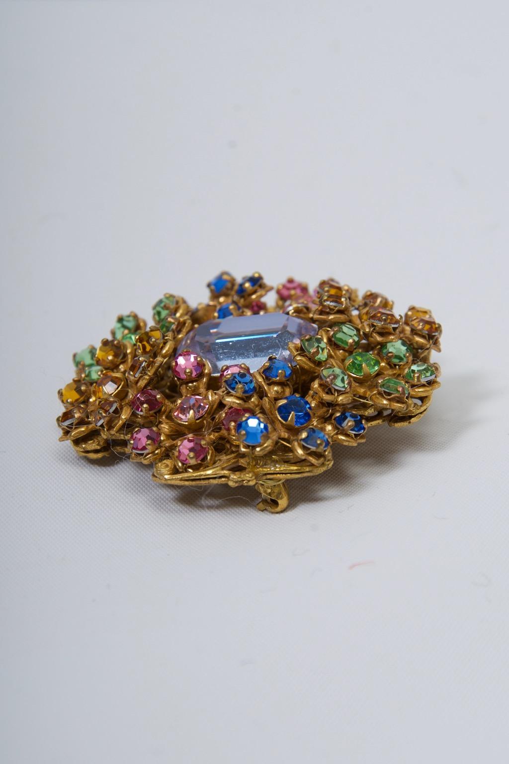 Vintage Miriam Haskell brooch, the pierced gold metal frame featuring sections of pastel crystals in pink, green, blue, and yellow centering a large emerald-shaped pale blue crystal. Oval plaque marked 
