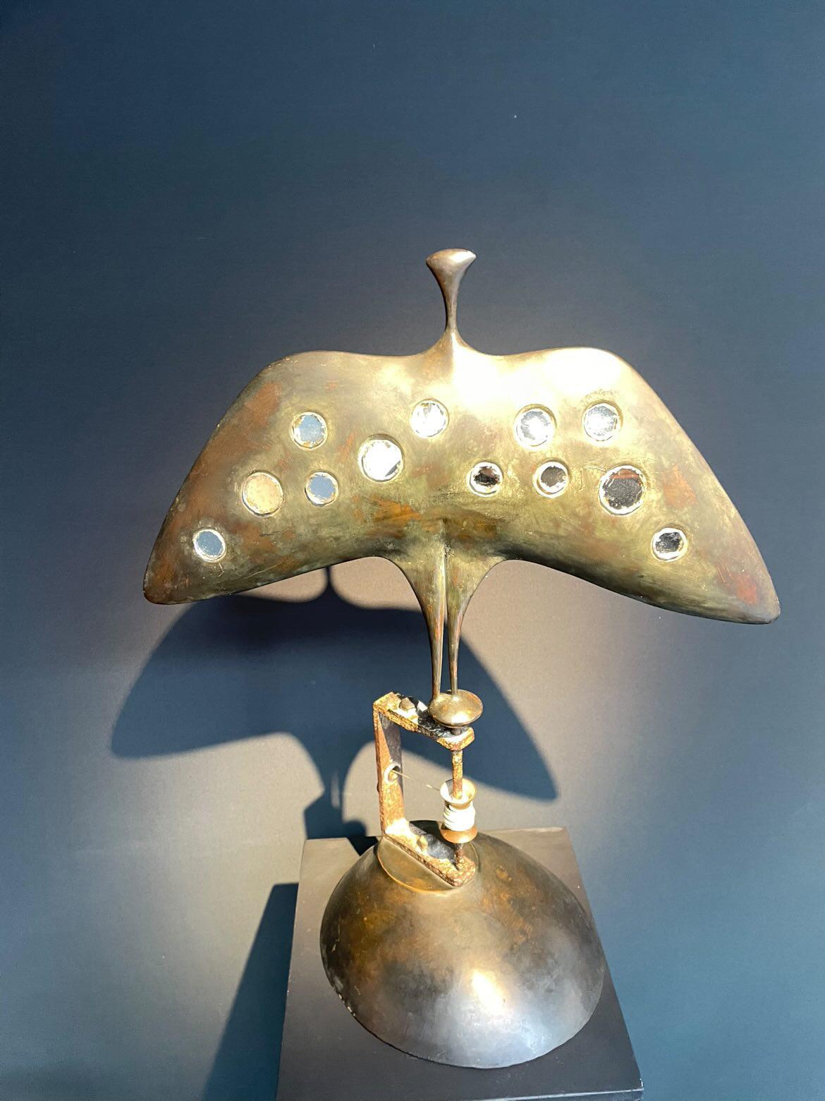 Cette sculpture cyclope qui fait penser à un oiseau s’actionne telle une toupie grâce à la ficelle qui s’enroule autour de l’avant du socle. Son corps est recouvert de plusieurs billes et d’yeux de verres colorés qui lorsque la sculpture tourne sur