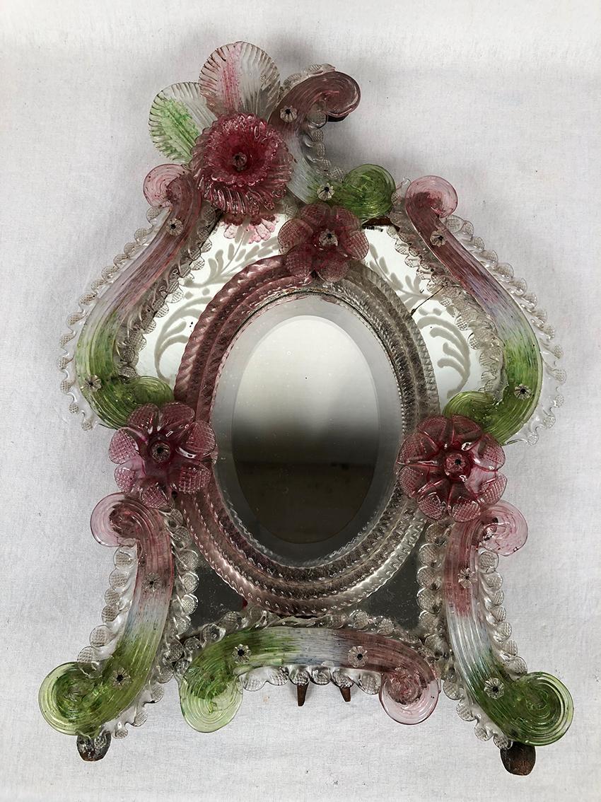 Miroir de table en verre coloré de Murano
travaillé à la pince,
miroir biseauté en médaillon entouré de miroirs gravés à décor de fleurs (un présente un fêle)
encadrés de moulures et de fleurs bicolores rose et vert.
Dos et support en bois de forme