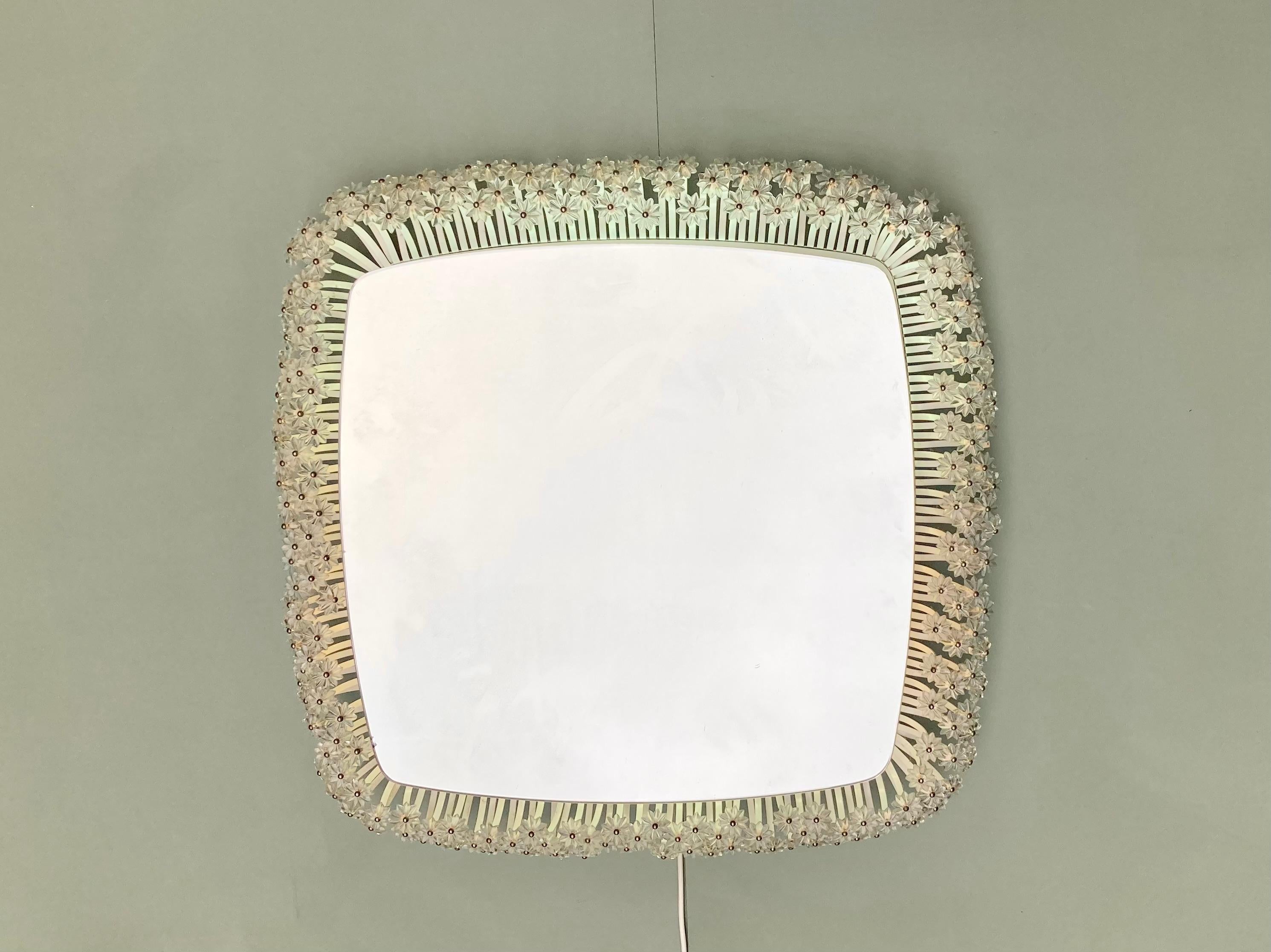 Miroir rétro-éclairé par Emil Stejnar pour Rupert Nikoll.
Le cadre du miroir est en métal laqué blanc terminé par des fleurs en verre.
La lumière est fonctionnelle, le miroir est parfait.