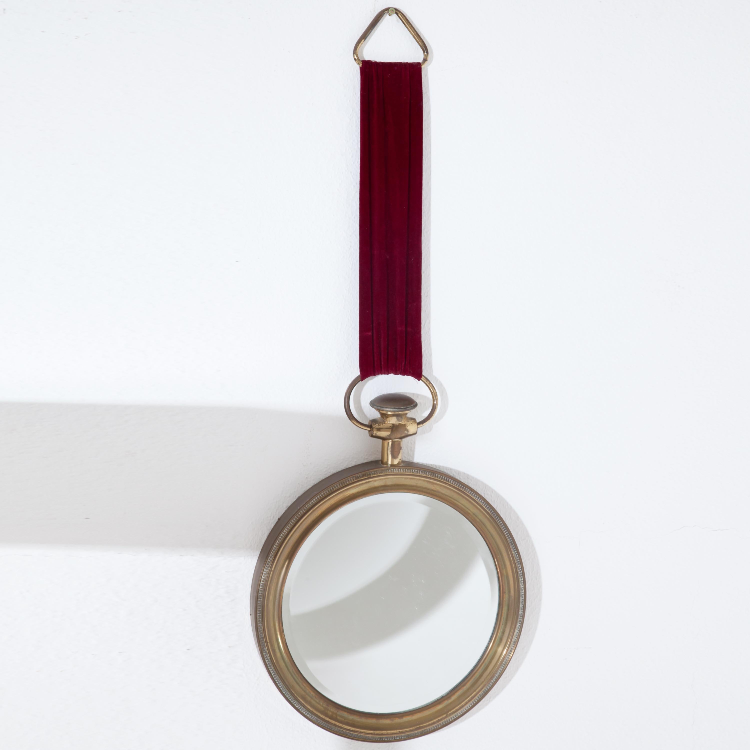 Round wall mirror by Piero Fornasetti in a profiled brass frame in the style of a pocket watch. The mirror with facetted mirror glass at the edge hangs on a long red velvet ribbon and has a triangle-shaped suspension.