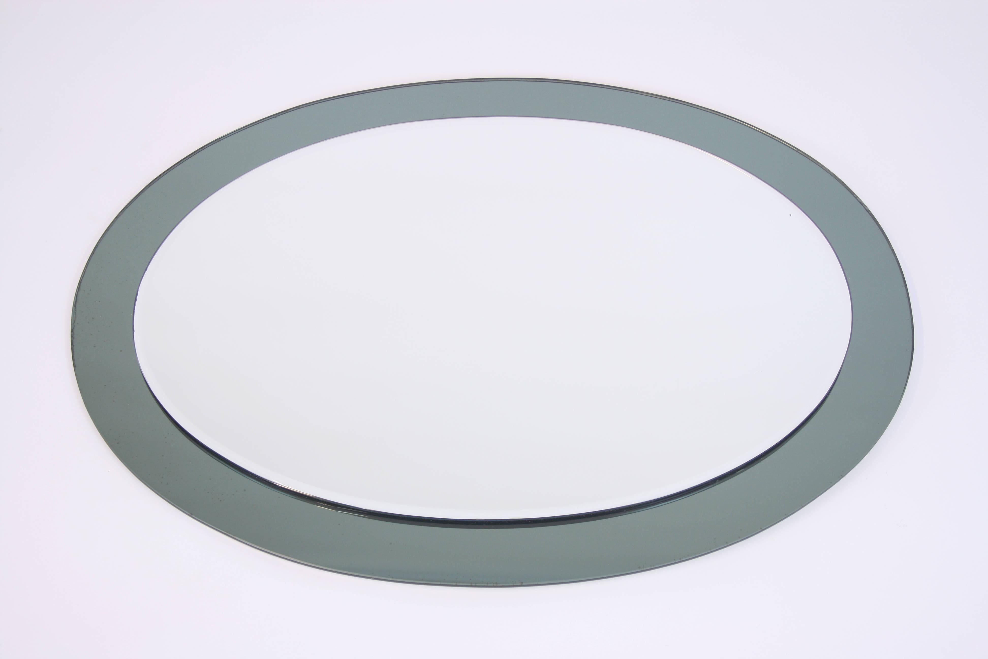 An oval curved mirror with a blue green shimmering frame, an original piece of the 1960s. Its softly honed and polished edges are reminiscent of products by Fontana Arte. An elegant eye-catcher for every spot. On the back is a barely visible mark of
