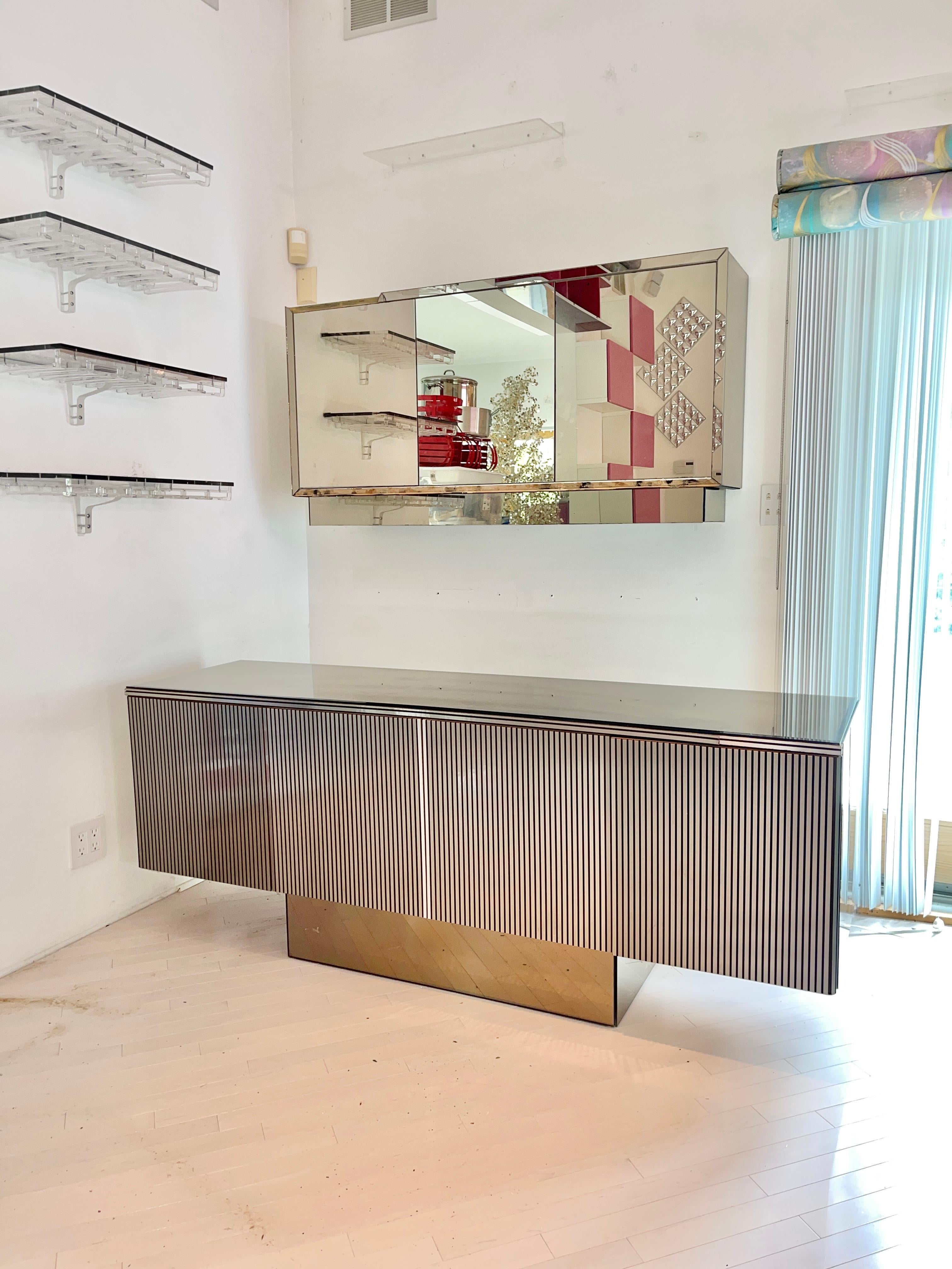 END OF SUMMER SALE

Four door floating sideboard clad in brushed aluminum ribbed laminate with smoked mirrored top and sides on recessed smoked mirrored platform base.

Includes a wall mounted mirrored three door back bar cabinet.  28x56x12. Can