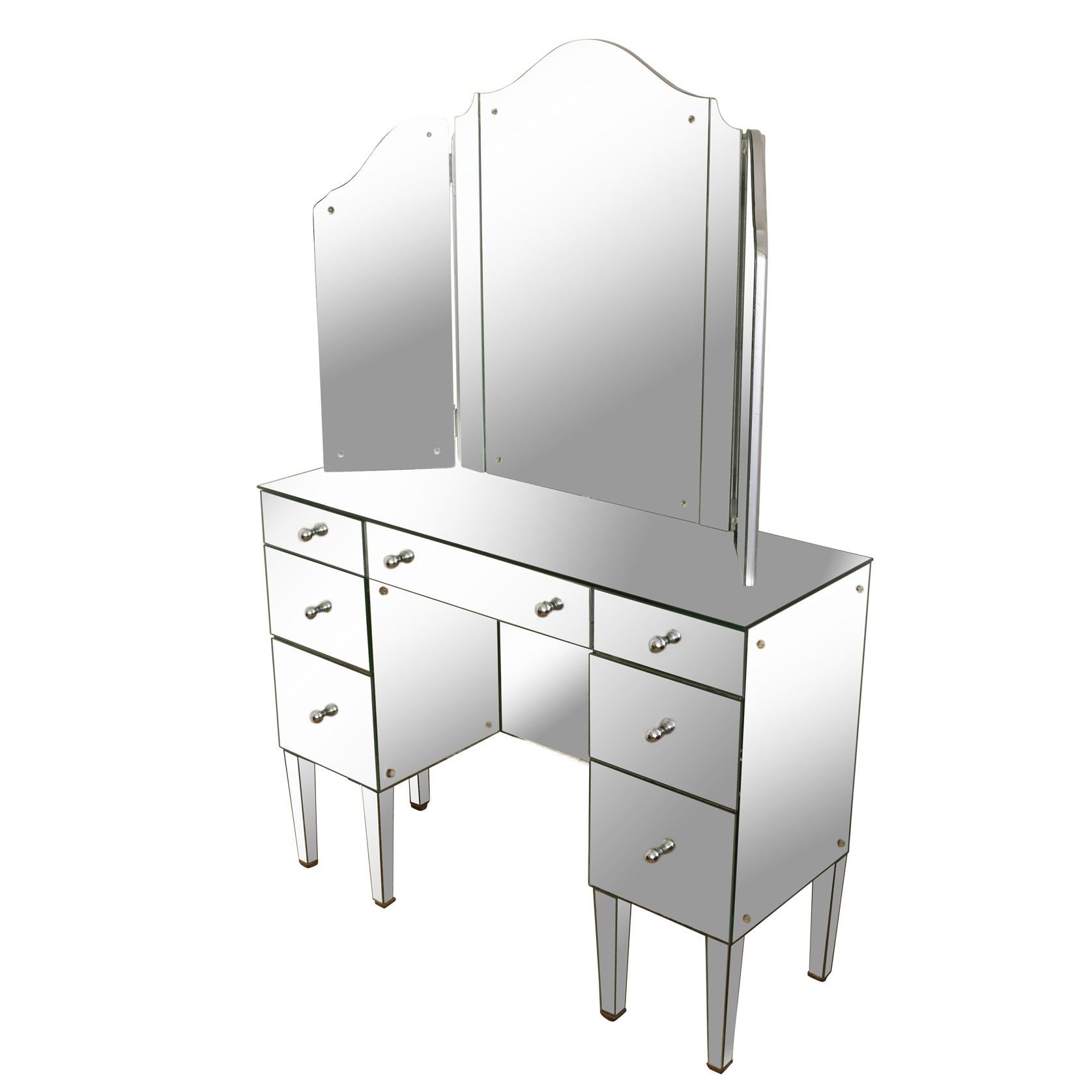 Une table de toilette vintage en miroir avec un miroir trifold attaché au plateau de la table de toilette.  Le meuble sous-lavabo à sept tiroirs est entièrement revêtu de miroir et les boutons de tiroirs sont ronds et en chrome poli.  Le miroir