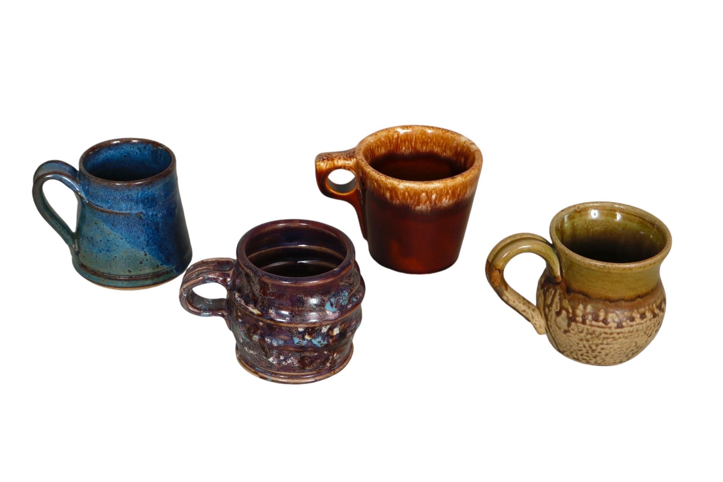 A set of four mismatched earthenware mugs with colorful drip glazes. One is a triangular shaped mug in a blue with green drip glaze, another is purple with a sculptural ribbed shape, signed underneath by the artist and ceramicist Ron Korczynski. A