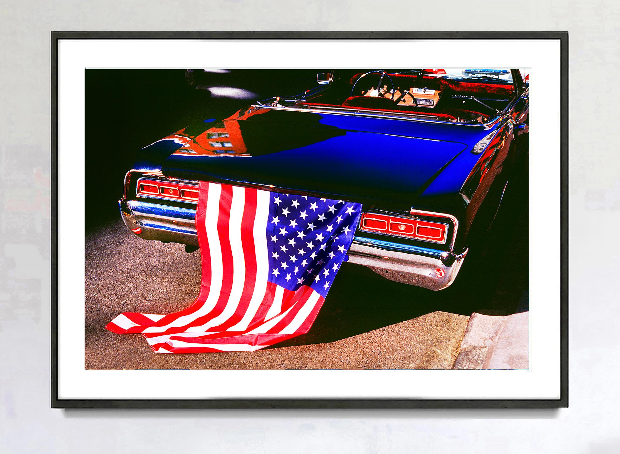 Quoi de plus patriotique qu'une décapotable rétro des années 70, bleu et rouge, avec un drapeau américain drapé sur le coffre ?  Le photographe de rue Mitchell Funk capture l'instant avec une lumière perçante, créant des couleurs vives et de longues
