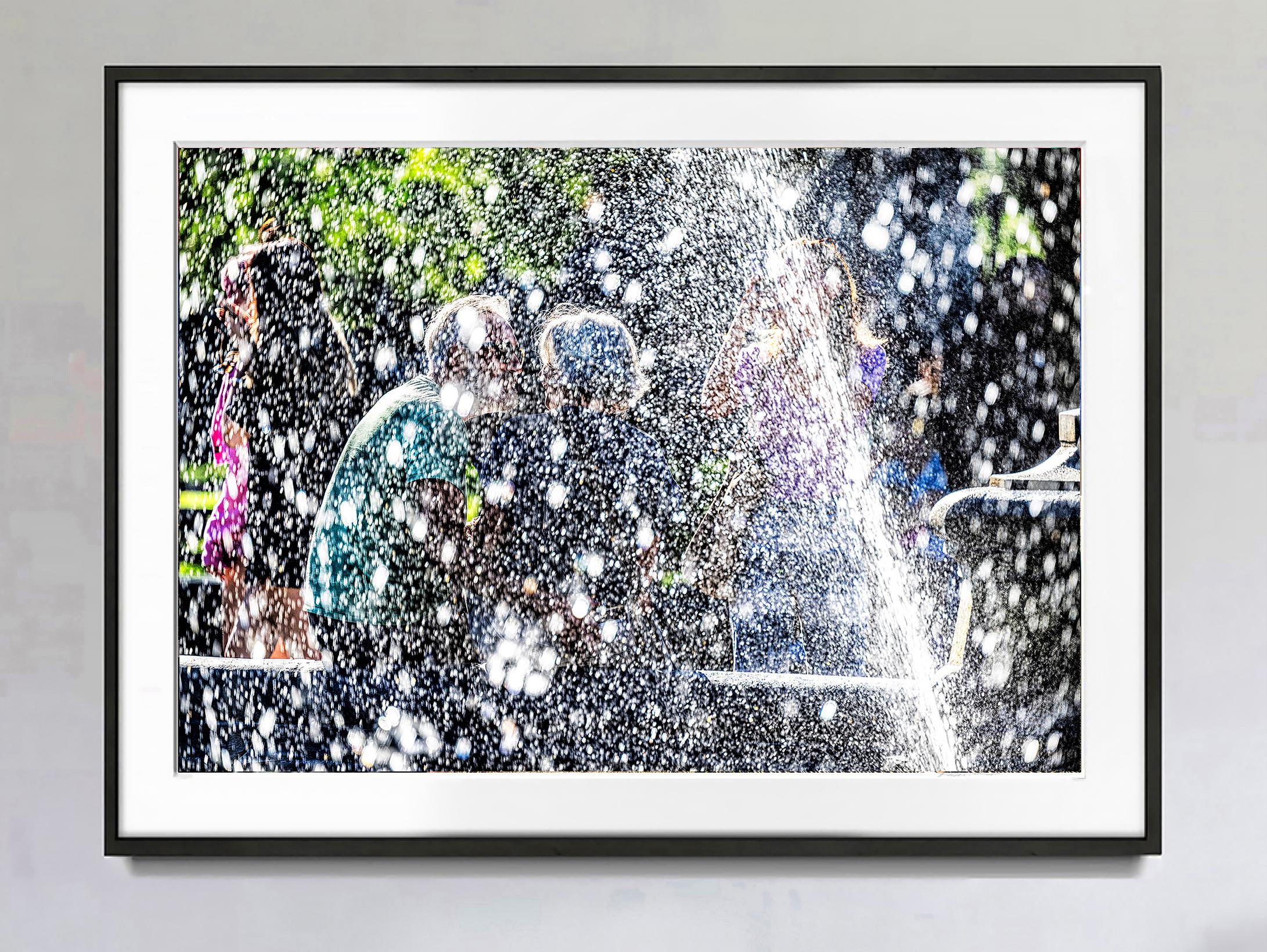 Blurry figures are vaguely seen through the dancing waters of the Washington Square Fountain in New York.  Raking light strikes the water streams, which results in dots and dashes of  Pointillist-like brushstrokes of light and color.  The immediate