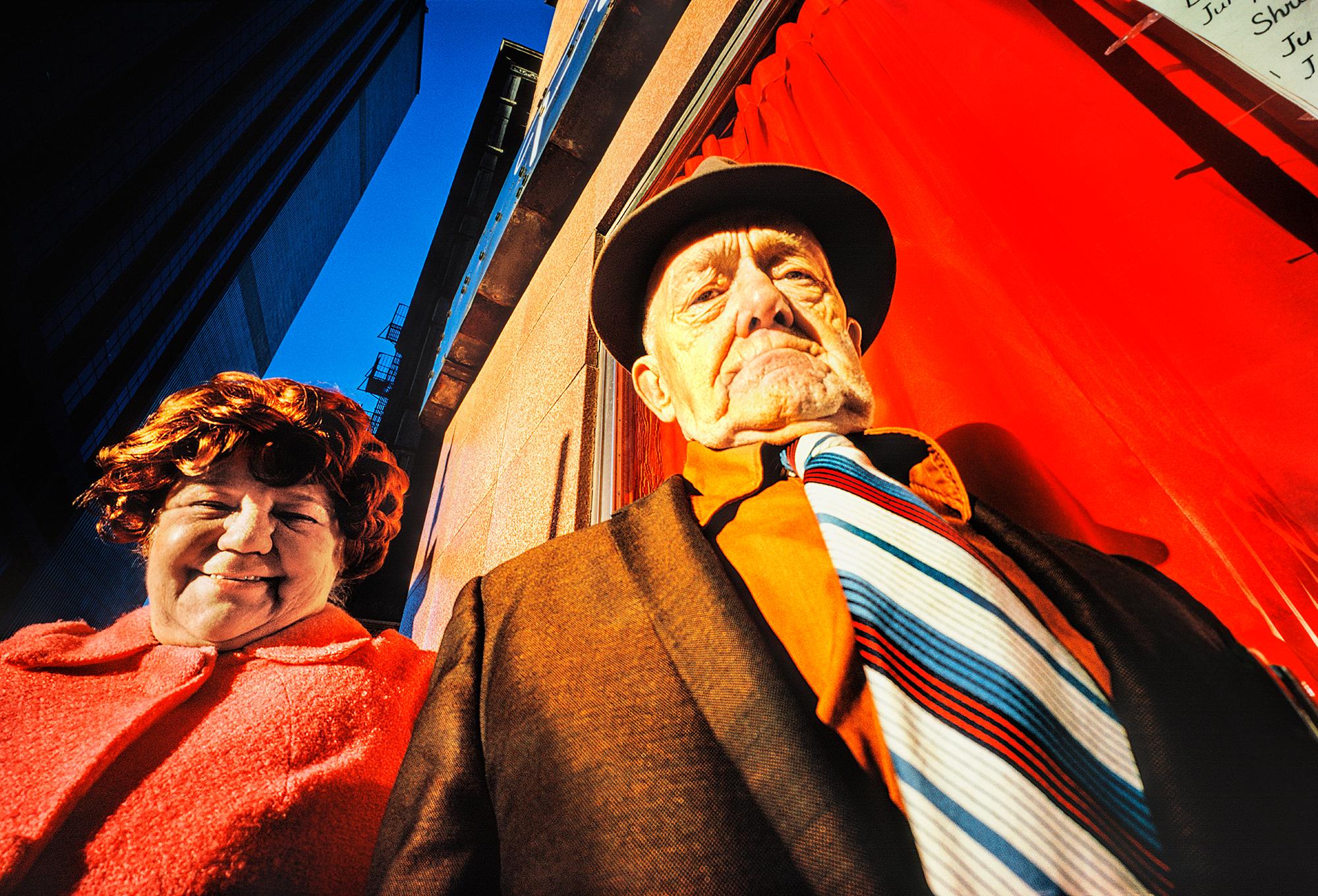 Mitchell Funk Color Photograph - Elderly Husband and Wife Group Street Portrait Against a Manhattan Red Wall