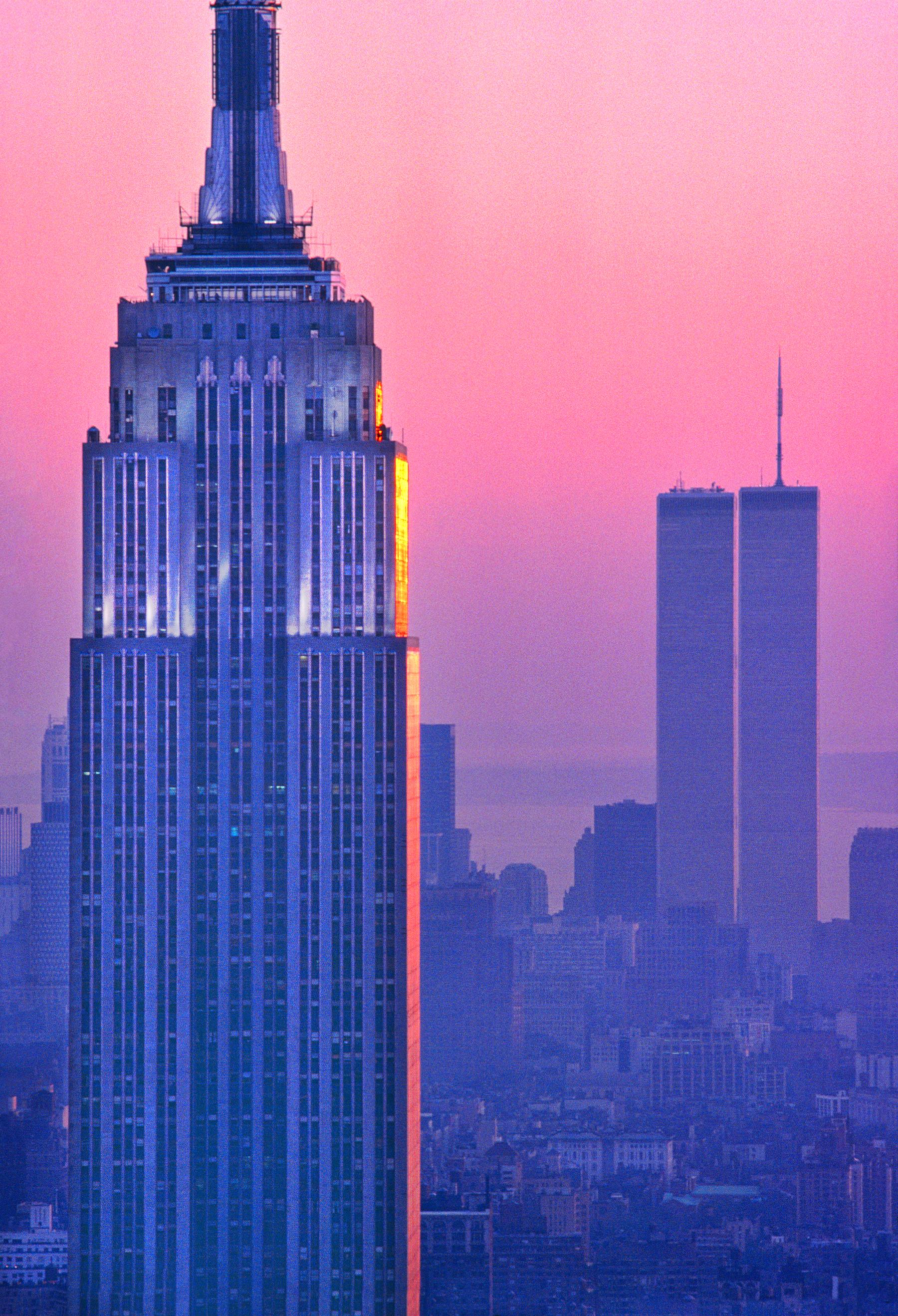 Zwei architektonische Ikonen von New York City werden vor einem magentafarbenen Himmel einander gegenübergestellt.  Das Empire State Building und die Zwillingstürme, aufgenommen vor einem Bauboom in Manhattan, bieten einen klaren und ungetrübten