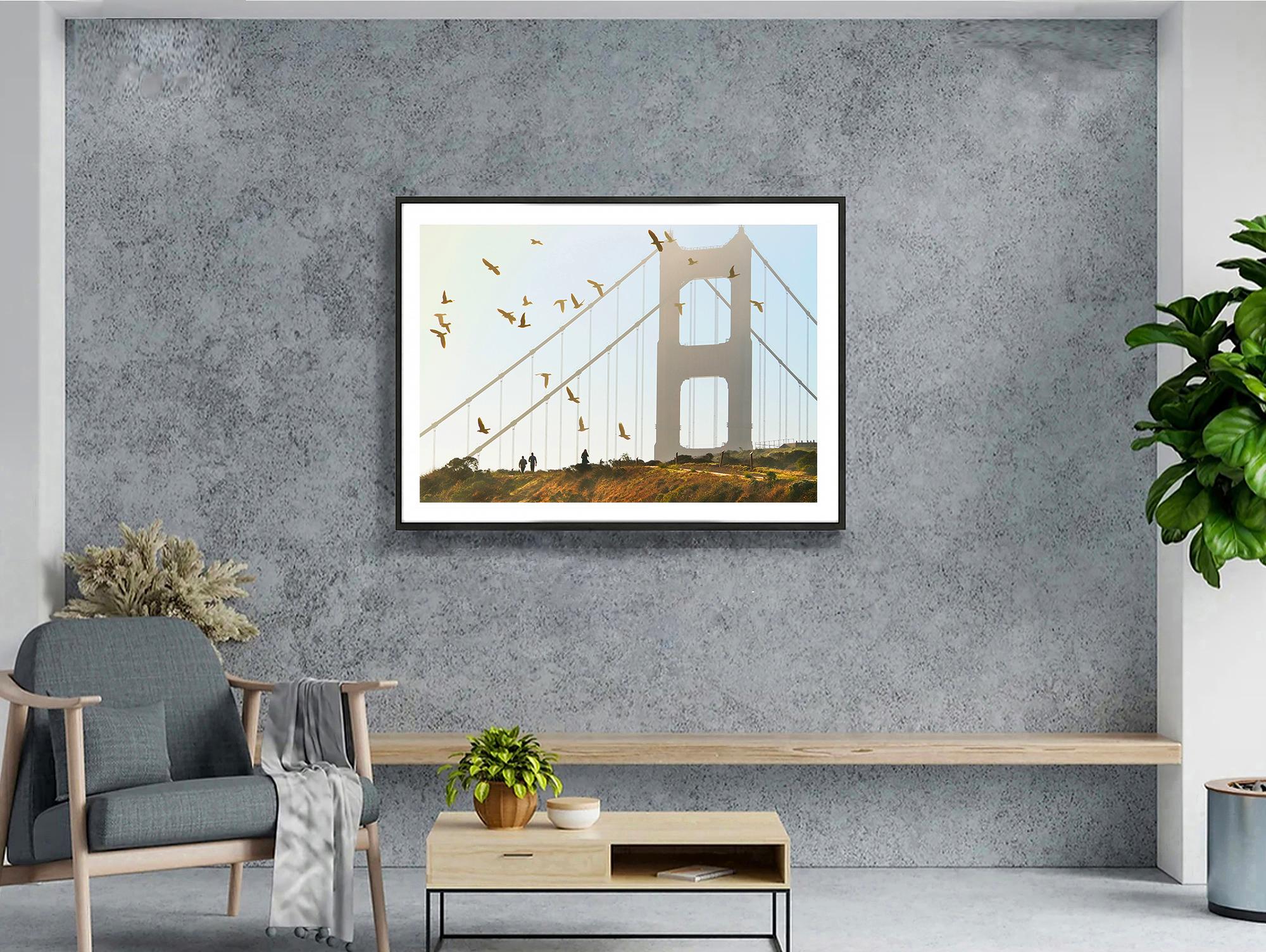 El arco semisilueteado del majestuoso puente Golden Gate es acariciado por los tonos suaves y cálidos de un sol poniente. En lo alto, una bandada de pájaros da vueltas como si rindiera homenaje al elevado edificio. En primer plano, tres personas dan