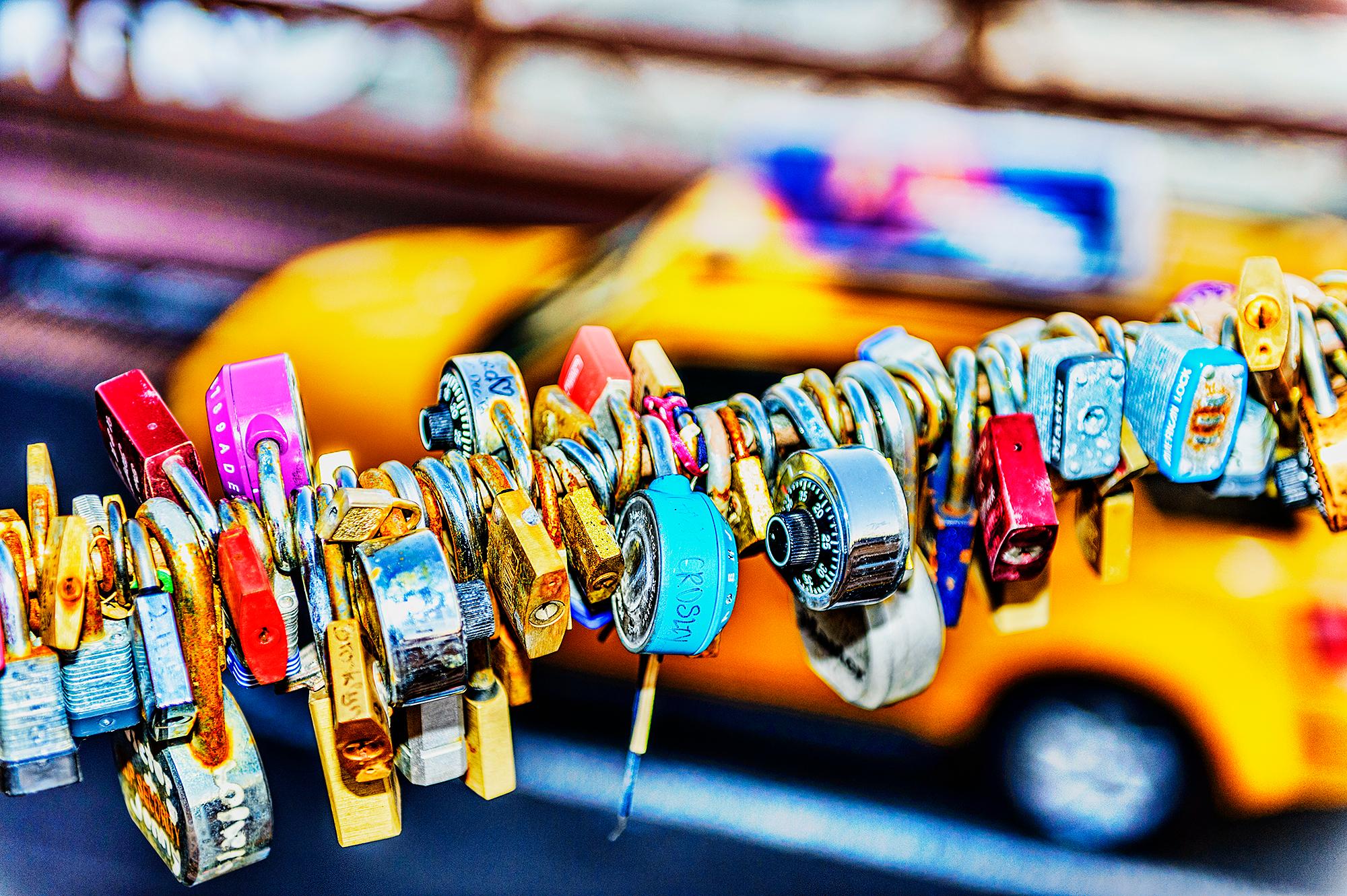 Colorful locks hanging from a wire above the Brooklyn Bridge are set against a blurred yellow 
New York City Taxi Cab.  They form an unexpected ambiguous abstract image composed of intersecting diagonal planes.   There is a great diversity of locks