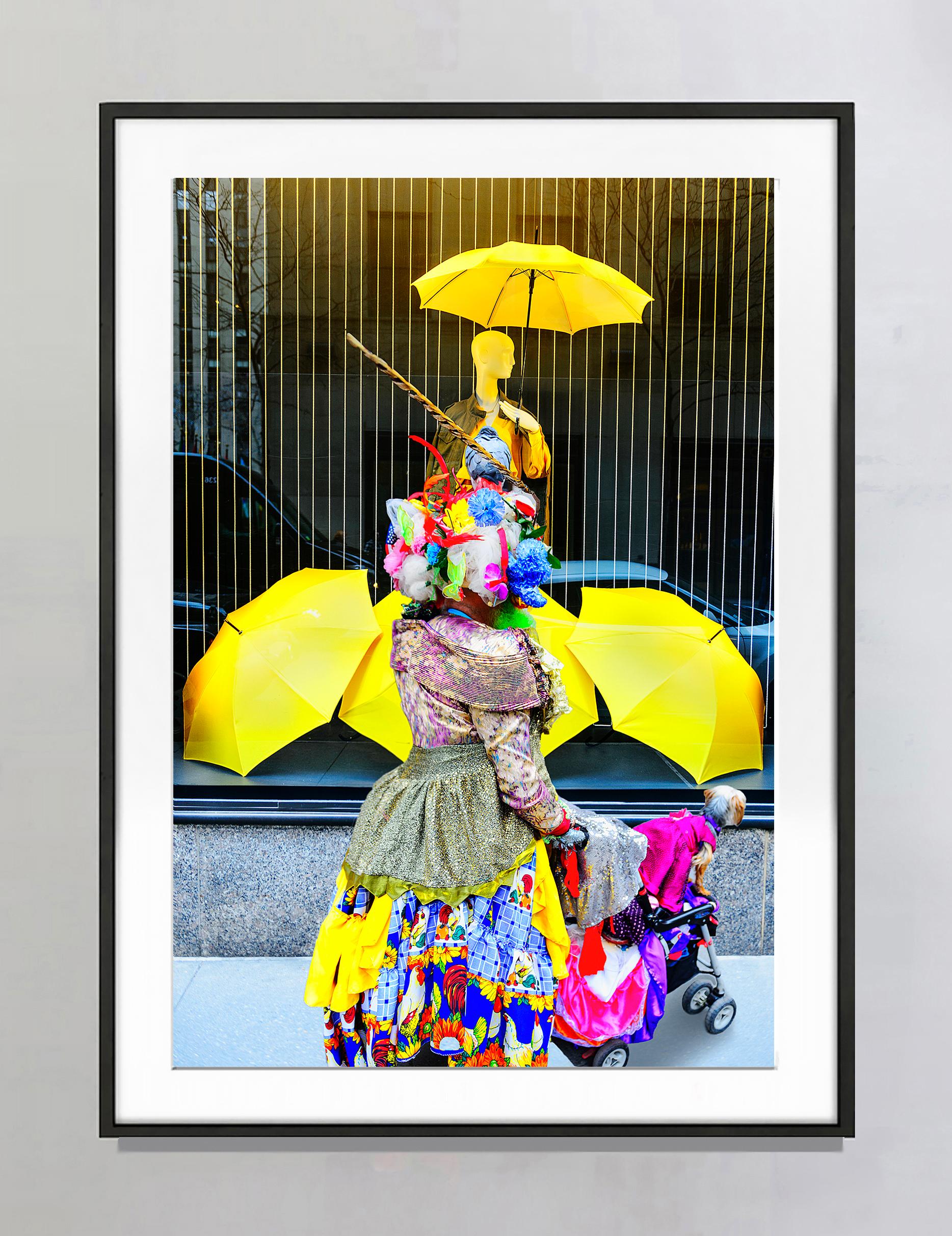 Street photograph of an eccentric woman with a brightly colored hat and colorful clothes pushing a baby carriage with a dog occupant. She stands in front of a window display with bright yellow umbrellas. The interaction between the colors of the