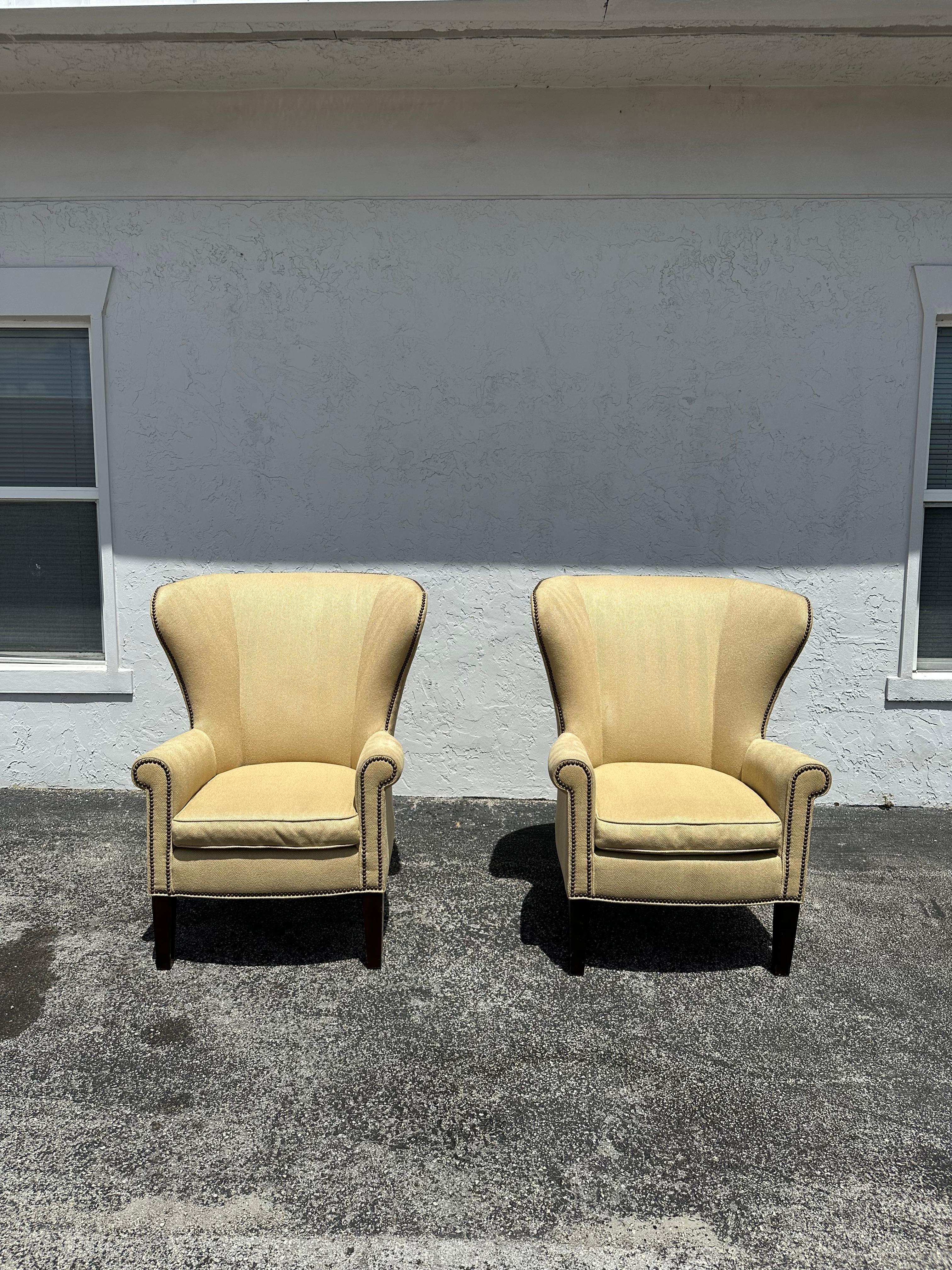 Pair of Wingback Chairs by Mitchell Gold + Bob Williams. The chairs are upholstered in a neutral chenille with a herringbone texture. The wood legs are square with a taper, with a black finish. Nailheads accent the front apron and perimeters of the