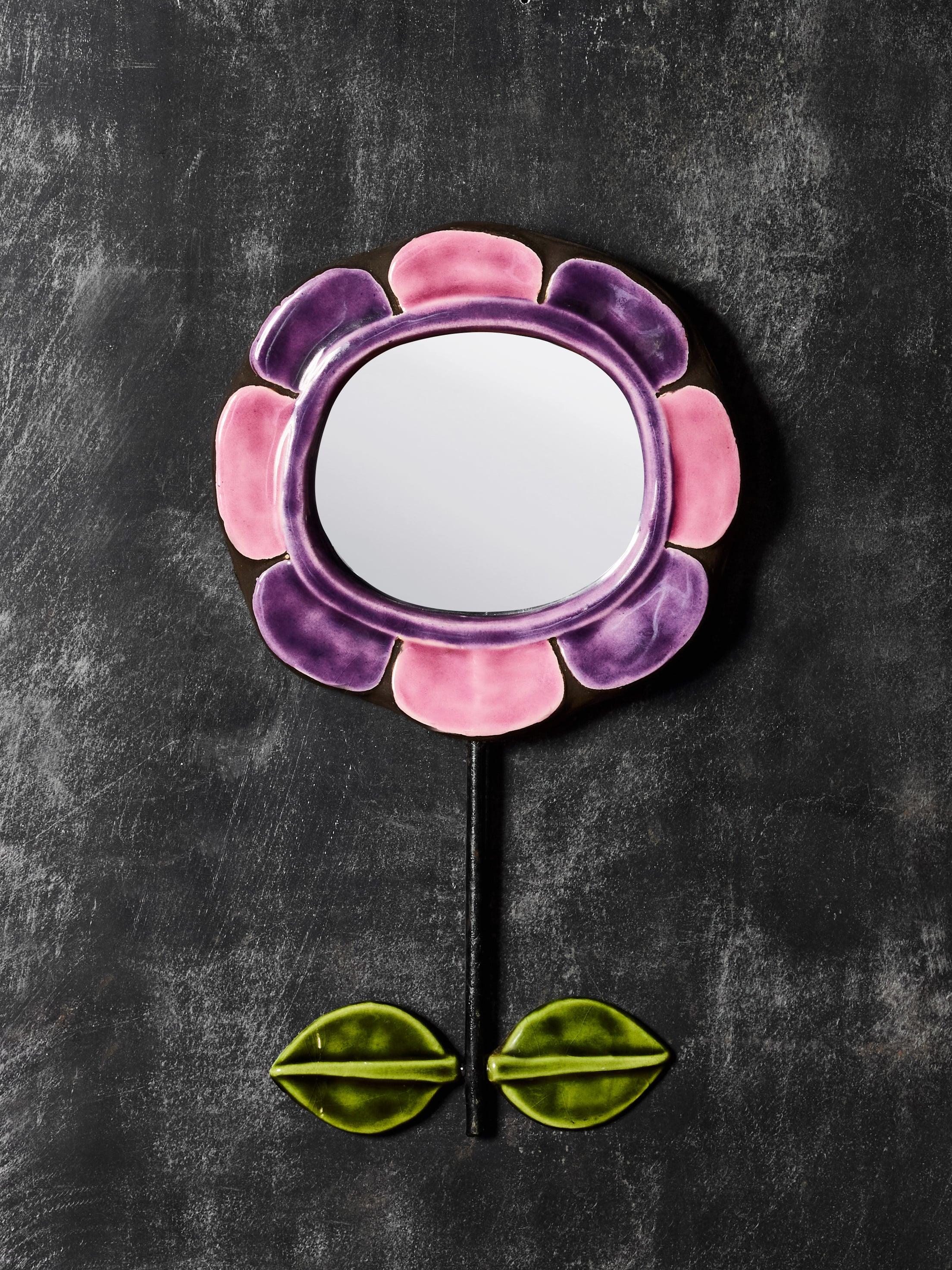 Charmant miroir en céramique de Mithé Espelt, fabriqué dans les années 1960 et en forme de fleur stylisée. Le cadre présente des éléments en forme de pétales émaillés de rose tendre et de violet profond, avec une tige noire élancée et deux feuilles