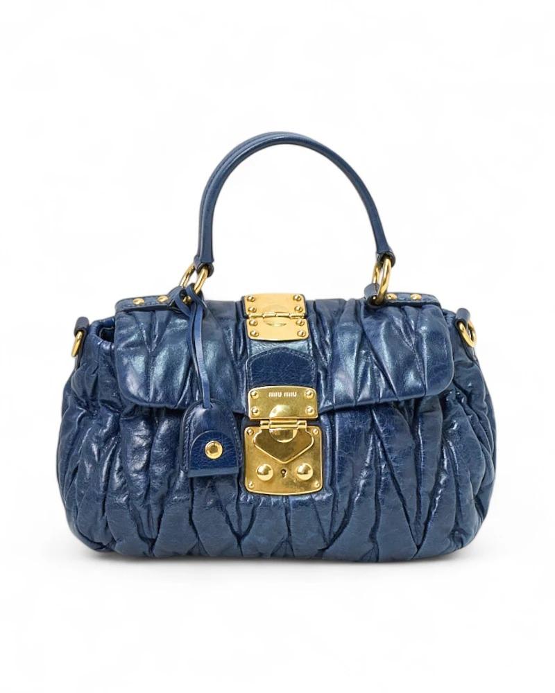 El bolso emblemático de Miu Miu, un bowling bag en tamaño pequeño, realizado en piel azul estriada con herrajes dorados. Equipado con una solapa frontal con cierre entrelazado y forrado interiormente en satén gris con un práctico bolsillo, el bolso