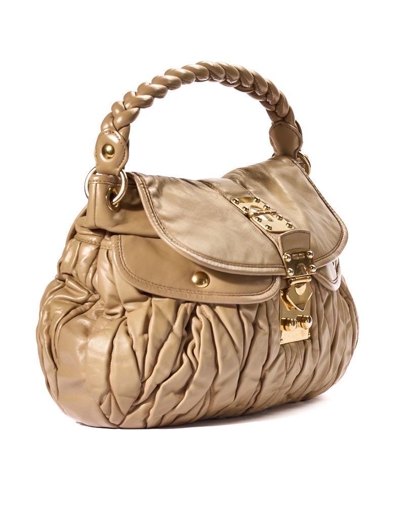 Borsa firmata Miu Miu, modello Coffer nella misura large, realizzata in pelle groffata beige con hardware dorato. Dotata di una patta frontale con chiusura ad incastro, internamente rivestita in tela beige con comoda tasca, la borsa è abbastanza