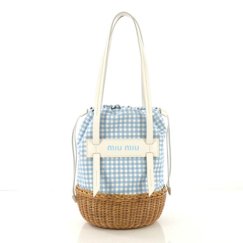 This Miu Miu Grace Bucket Bag Gingham Print Canvas and Wicker, crafted from blue and white gingham print canvas and brown wicker, features flat leather handles and logo across the front. Its drawstring closure opens to a blue canvas interior with