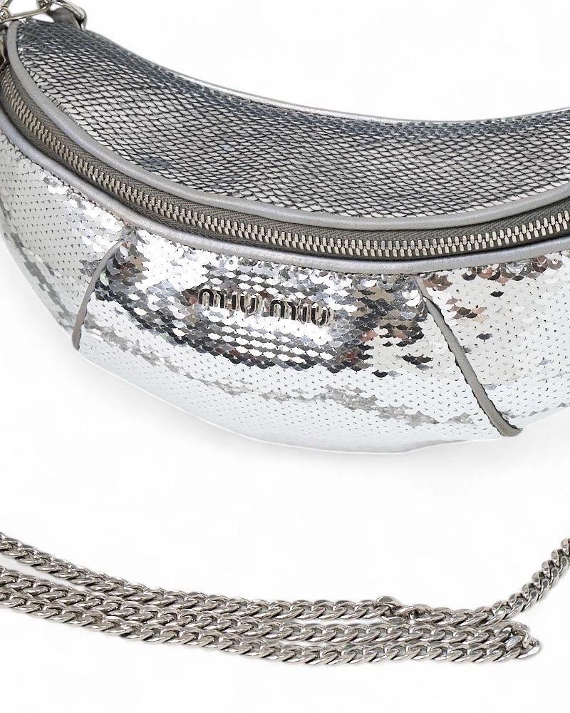 Marsupio firmato Miu Miu, misura small, realizzato in pelle argento arricchita da paillettes e hardware tono su tono. Dotato di una chiusura semicircolare a zip, internamente rivestito in tessuto nero, capiente per l’essenziale. Munito di una