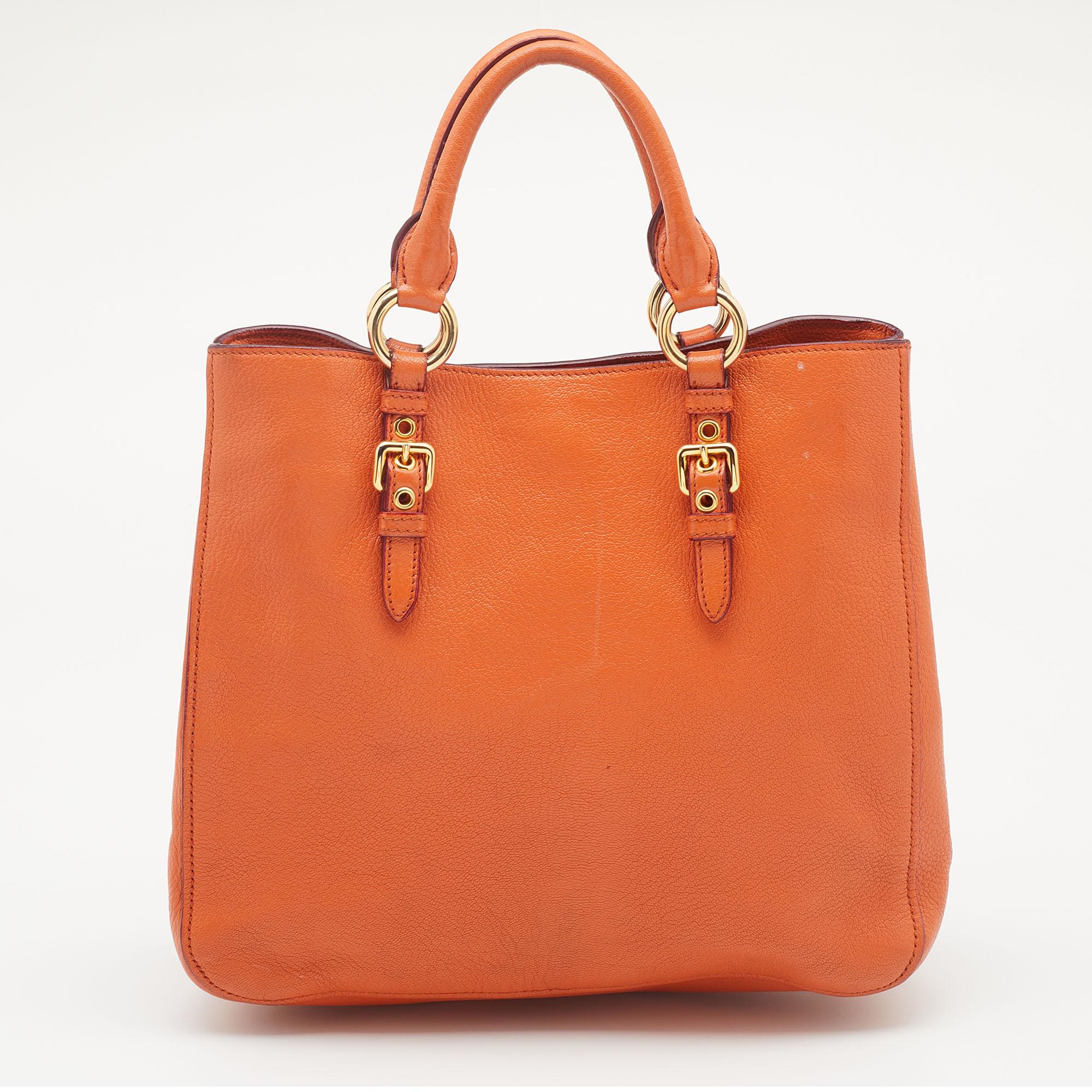 This Madras tote from the House of Miu Miu is attractive, sturdy, and practical for everyday use. It is crafted using orange leather on the exterior. It showcases two handles, gold-toned hardware, and a satin-lined interior. It is provided with an