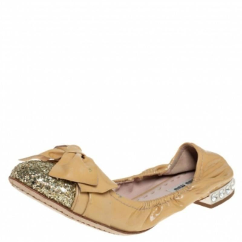 Created by Miu Miu, these scrunch ballet flats are a staple style every shoe collection needs. Constructed using patent leather, they feature glitter cap toes, bow details, and low heels embellished with crystals. The beige & gold shades add to the