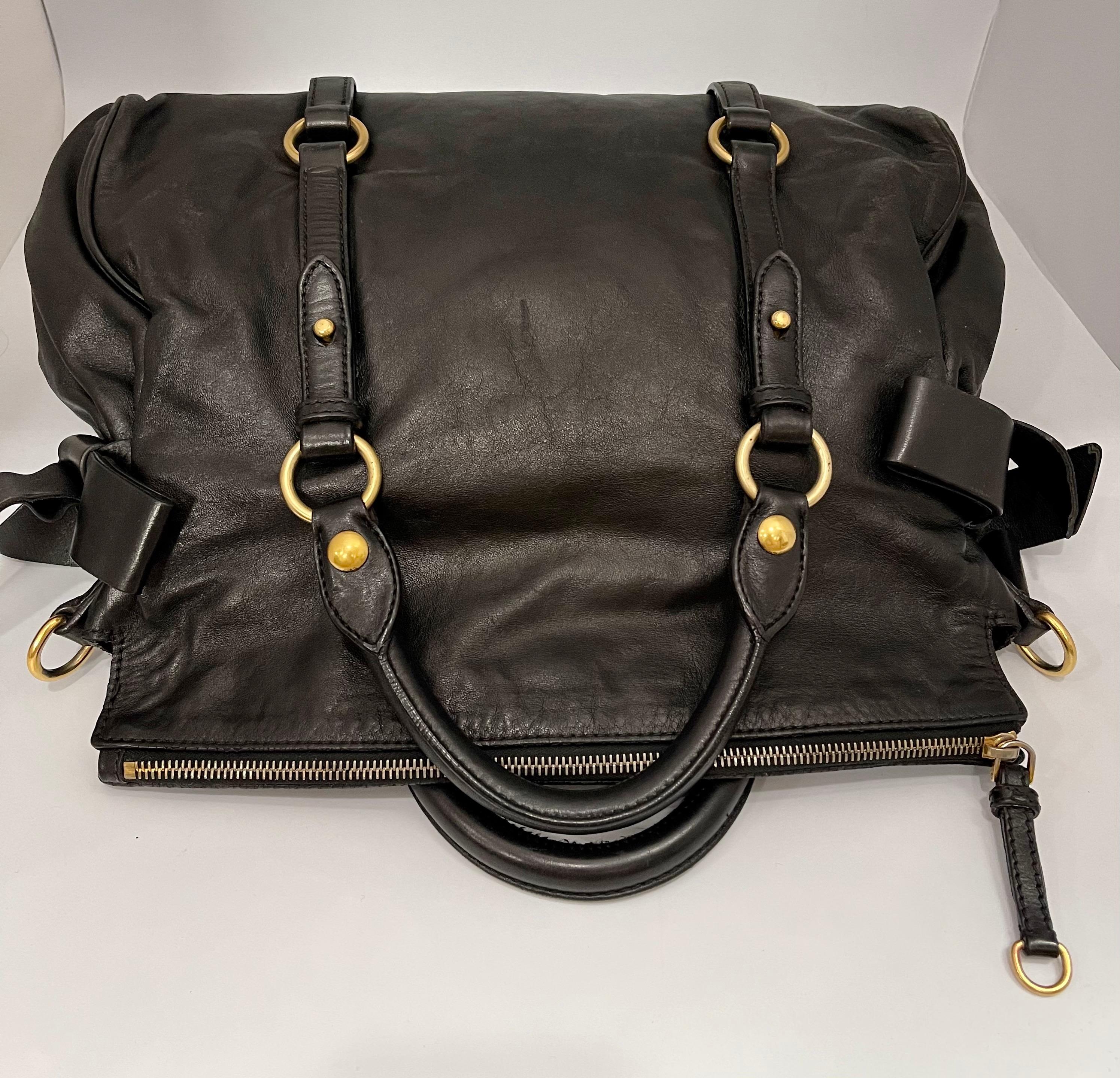Polished calfskin leather in black with large leather bows on the sides. Rolled leather top handles, an optional shoulder strap, belt embellishment around the girth, and polished brass hardware including large hoop links. The fold-over zip top opens