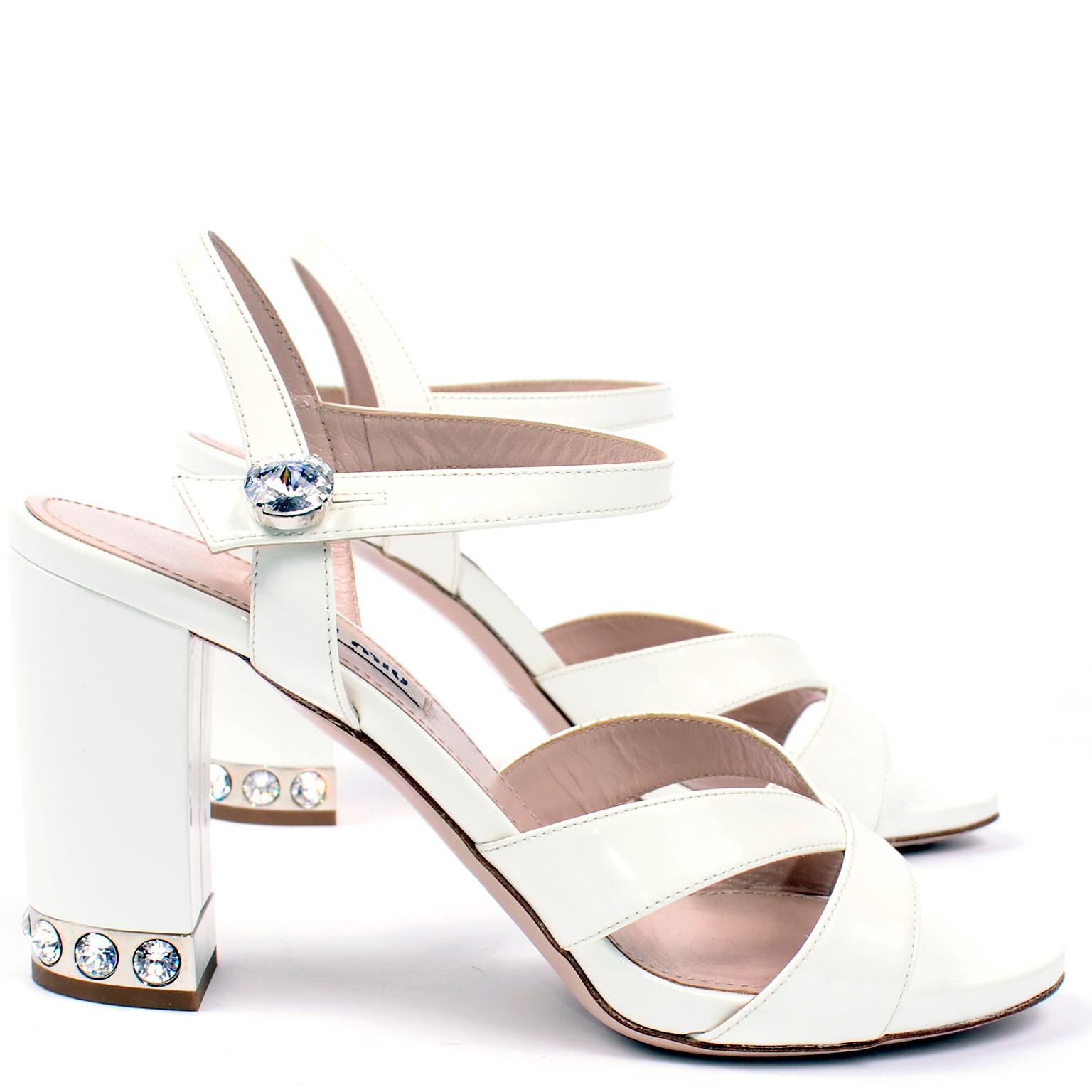 These are white leather strappy heeled open toe shoes from Miu Miu. There is a single ankle strap secured by a rhinestone button and the base of the heel has a silver tone plate with the same rhinestones around the heels. The tops of the shoes have