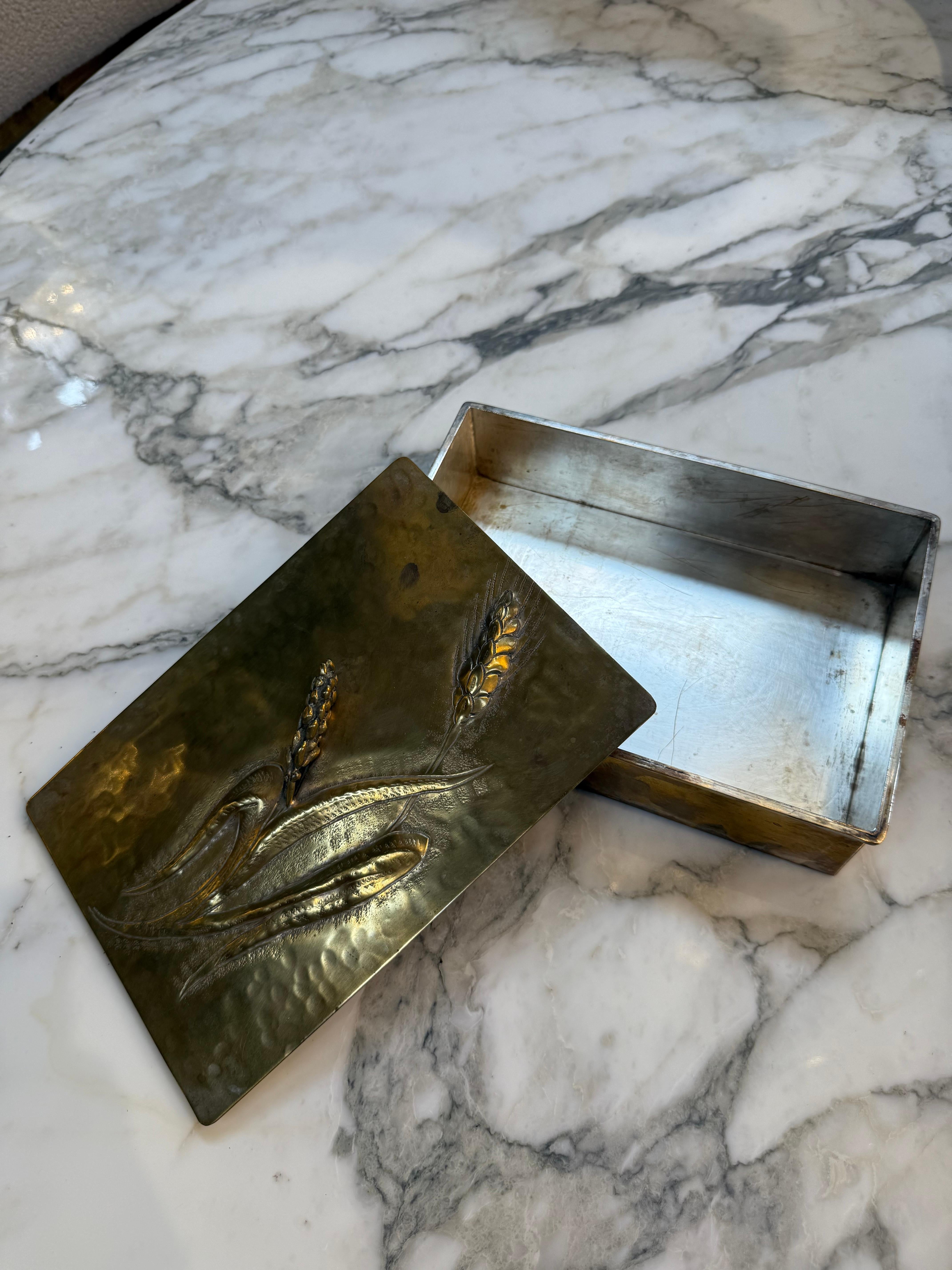 Questa raffinata scatola rettangolare di metà secolo presenta un elegante motivo floreale a intarsio realizzato con metalli contrastanti: fiori e foglie di rame caldo incastonati in un pannello di ottone lucido, delimitato da un fresco nichel