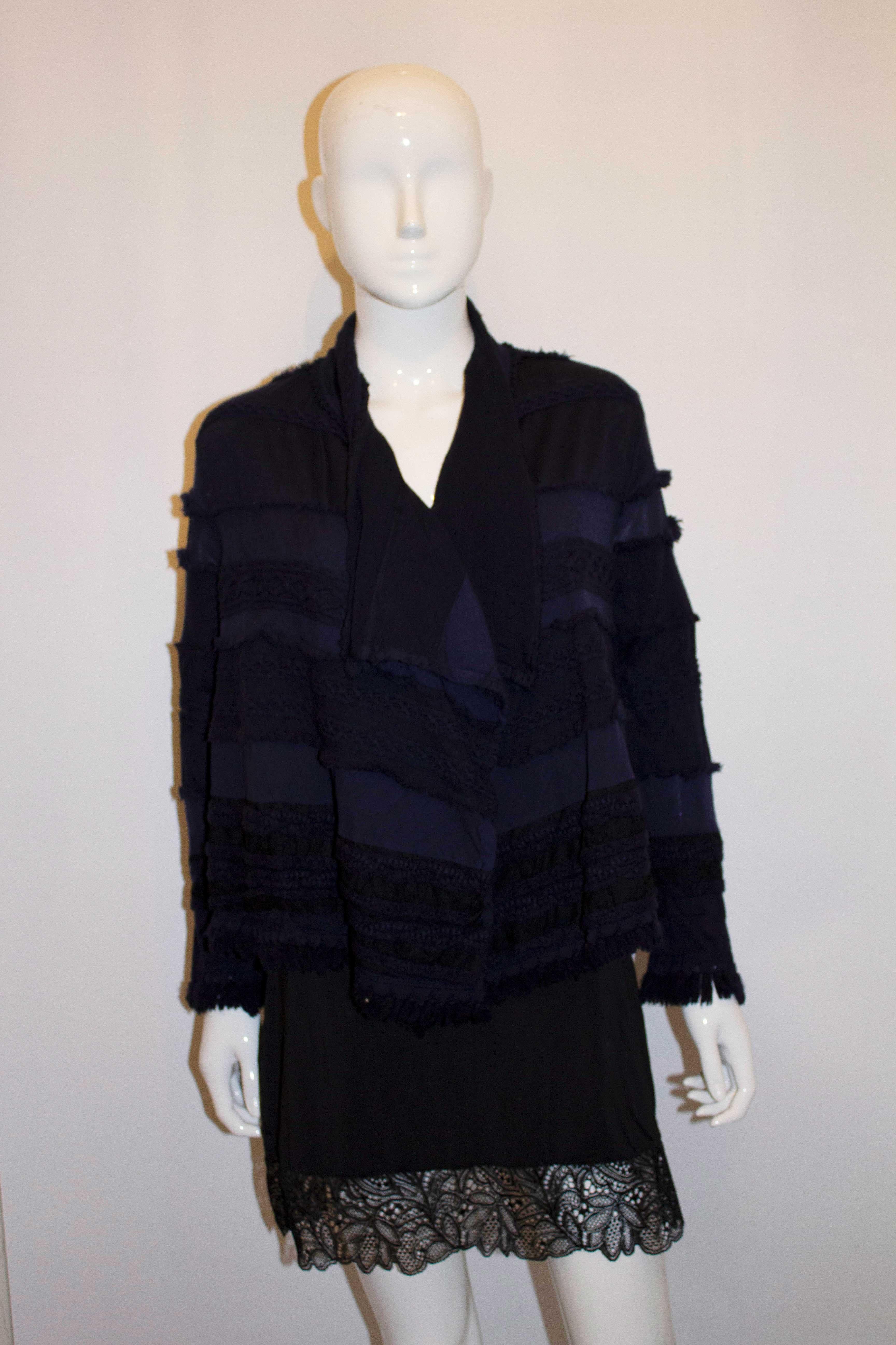 Une veste en laine facile à porter de la ligne Hatat de Miyake. Les manches et l'ourlet sont garnis de franges et le tissu est orné de galons et d'autres décorations. La veste est de couleur bleu encre foncé/noir, et n'est pas doublée. Taille 2